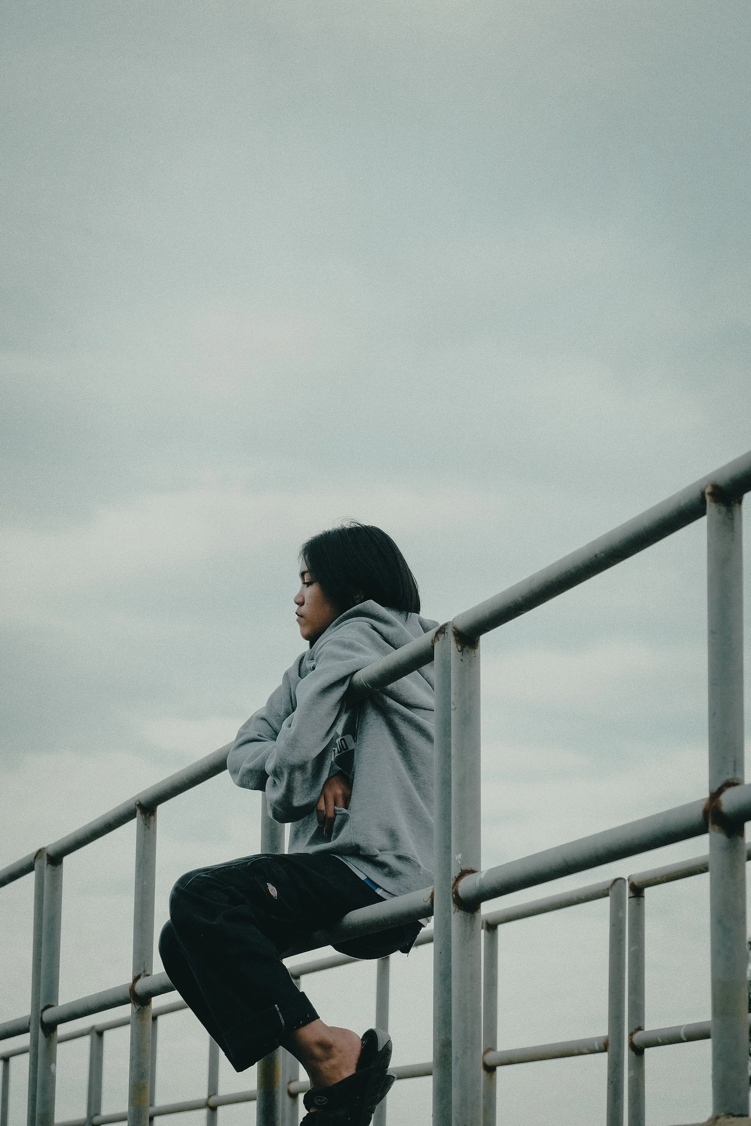 A person with dark hair sitting on a railing outdoors, wearing a gray hoodie and black pants, with an overcast sky in the background.