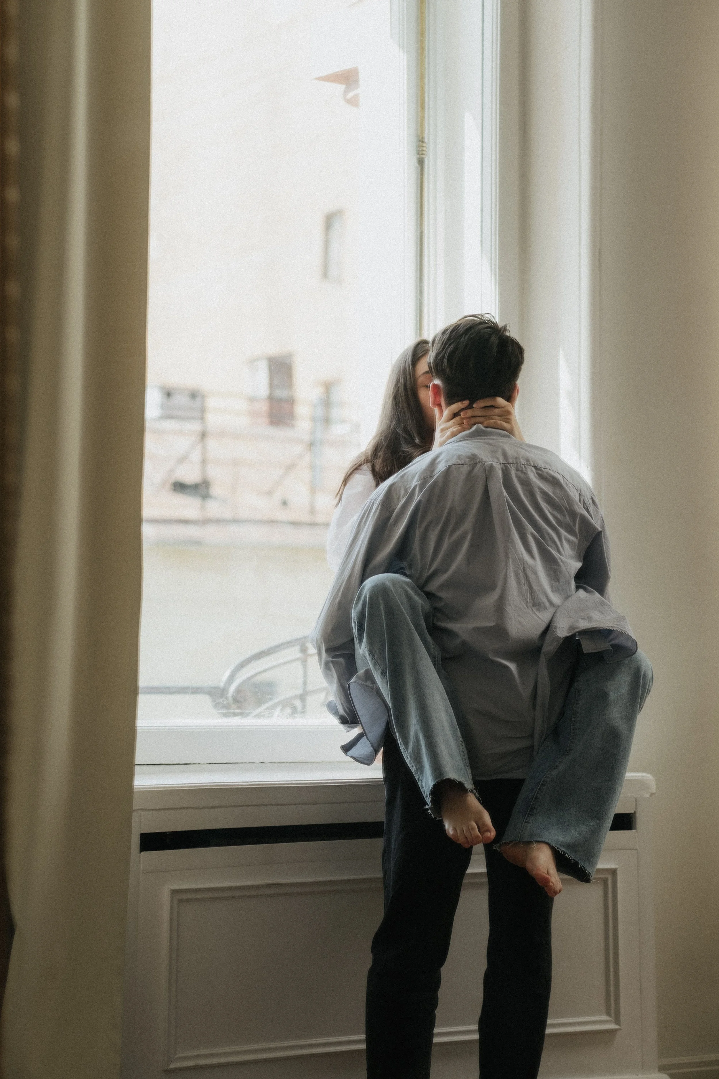 A couple kissing against a windowsill, with a cityscape seen through the window.