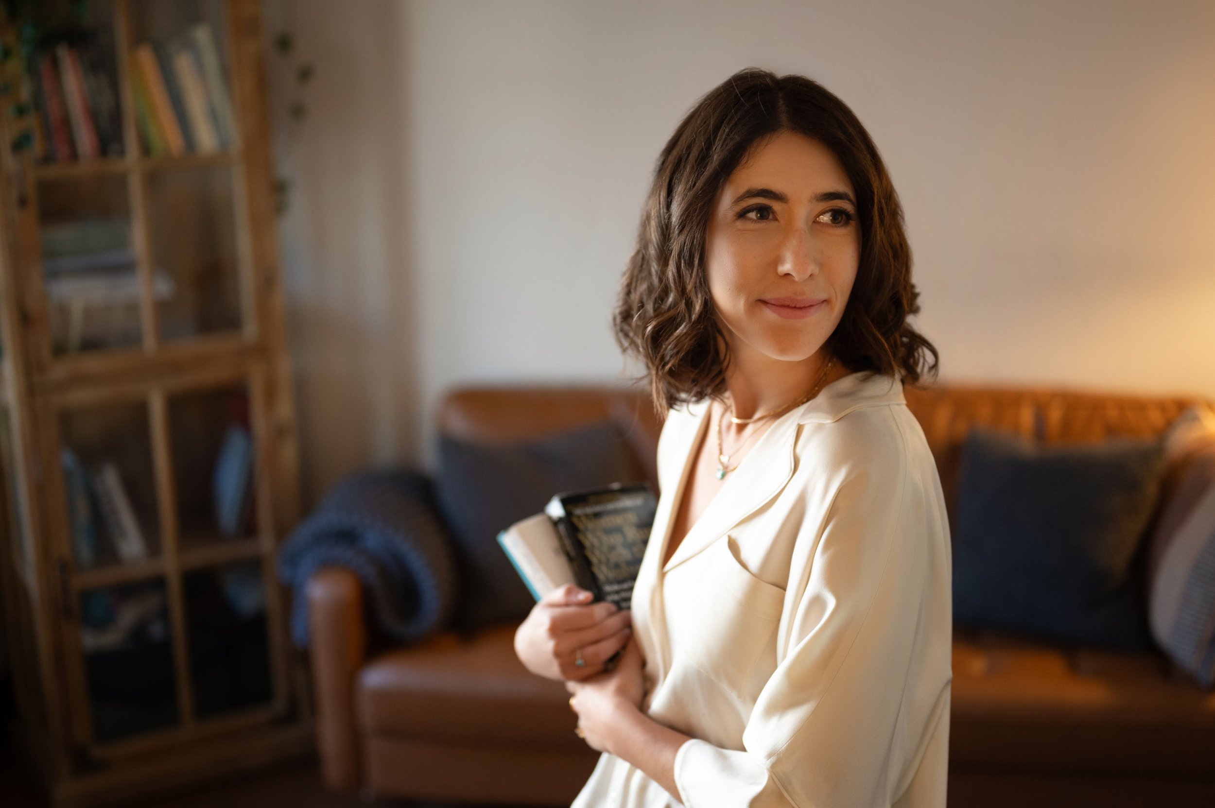 Deb Altman, LMFT, standing in a living room holding a book and a notebook, looking to her right with a slight smile.