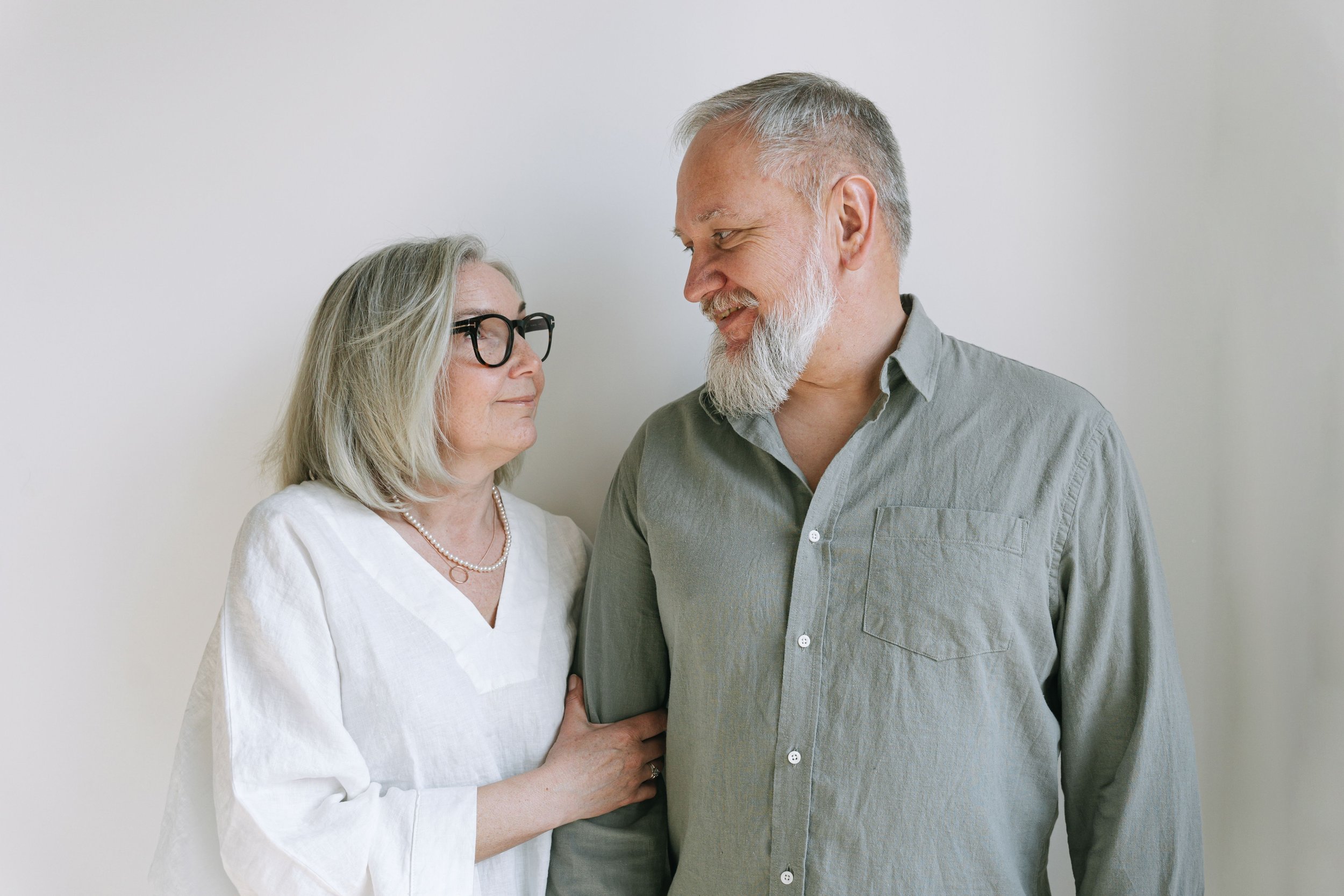 An elderly woman with gray hair, wearing glasses and a white blouse, looking lovingly at a middle-aged man with gray hair and a beard, wearing a gray shirt, against a plain white background.