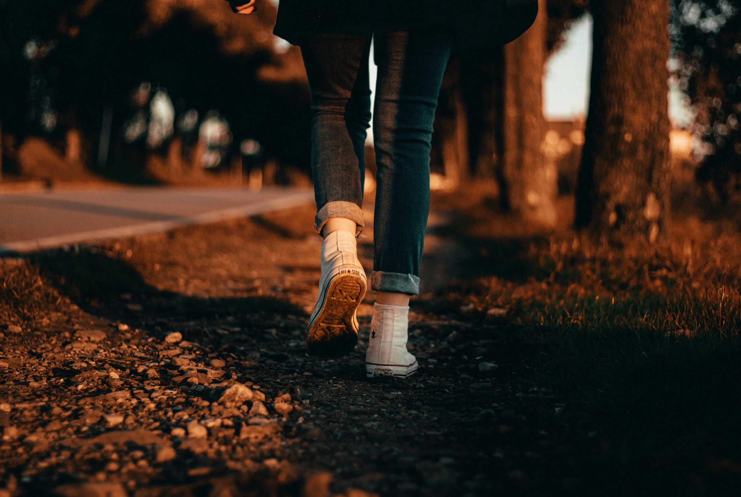 People walking outdoors on a gravel path during sunset, wearing jeans and sneakers.