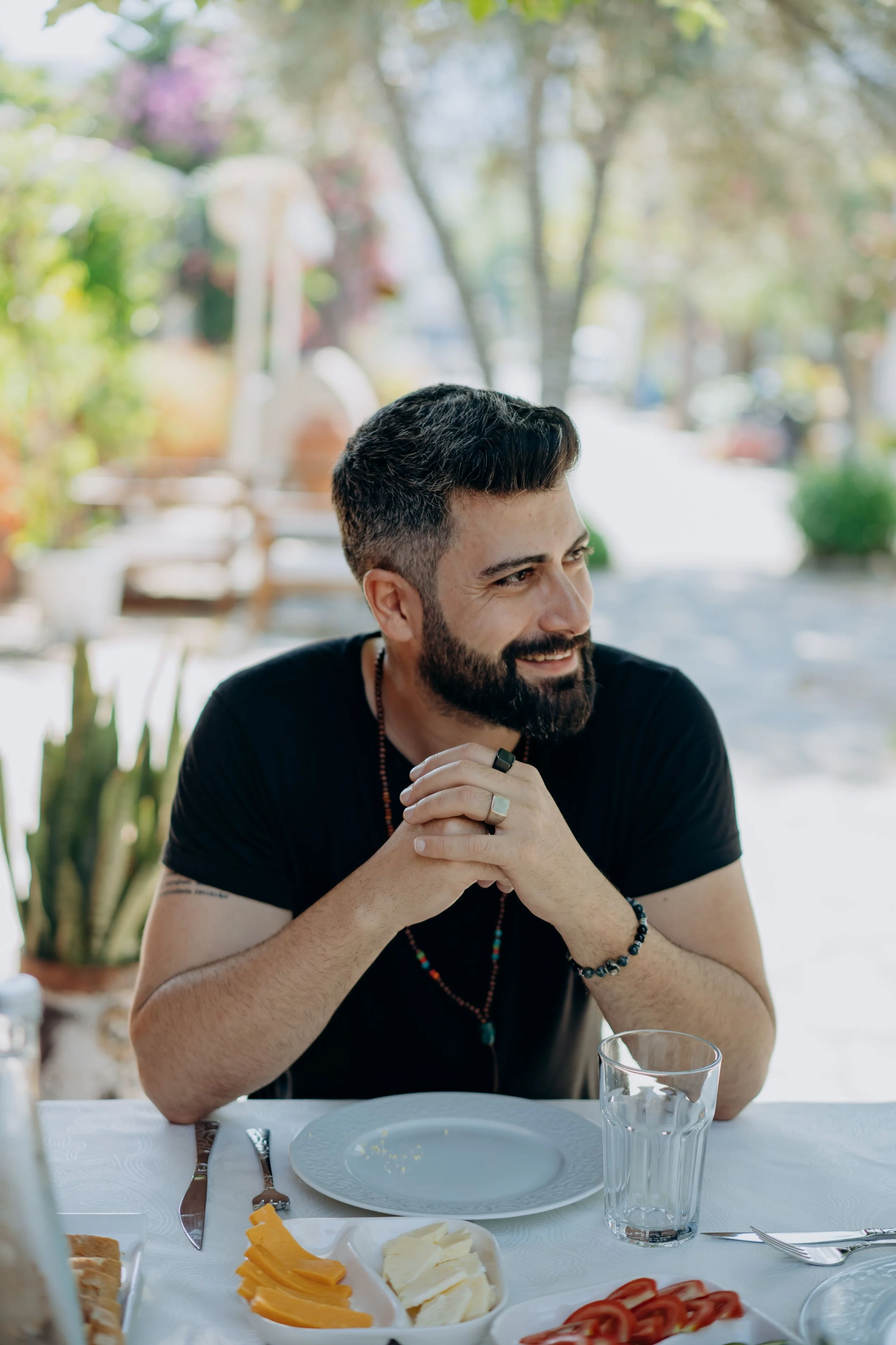 A man with dark hair and a beard smiling while sitting at a table outdoors, with food and drinks on the table, and trees in the background.