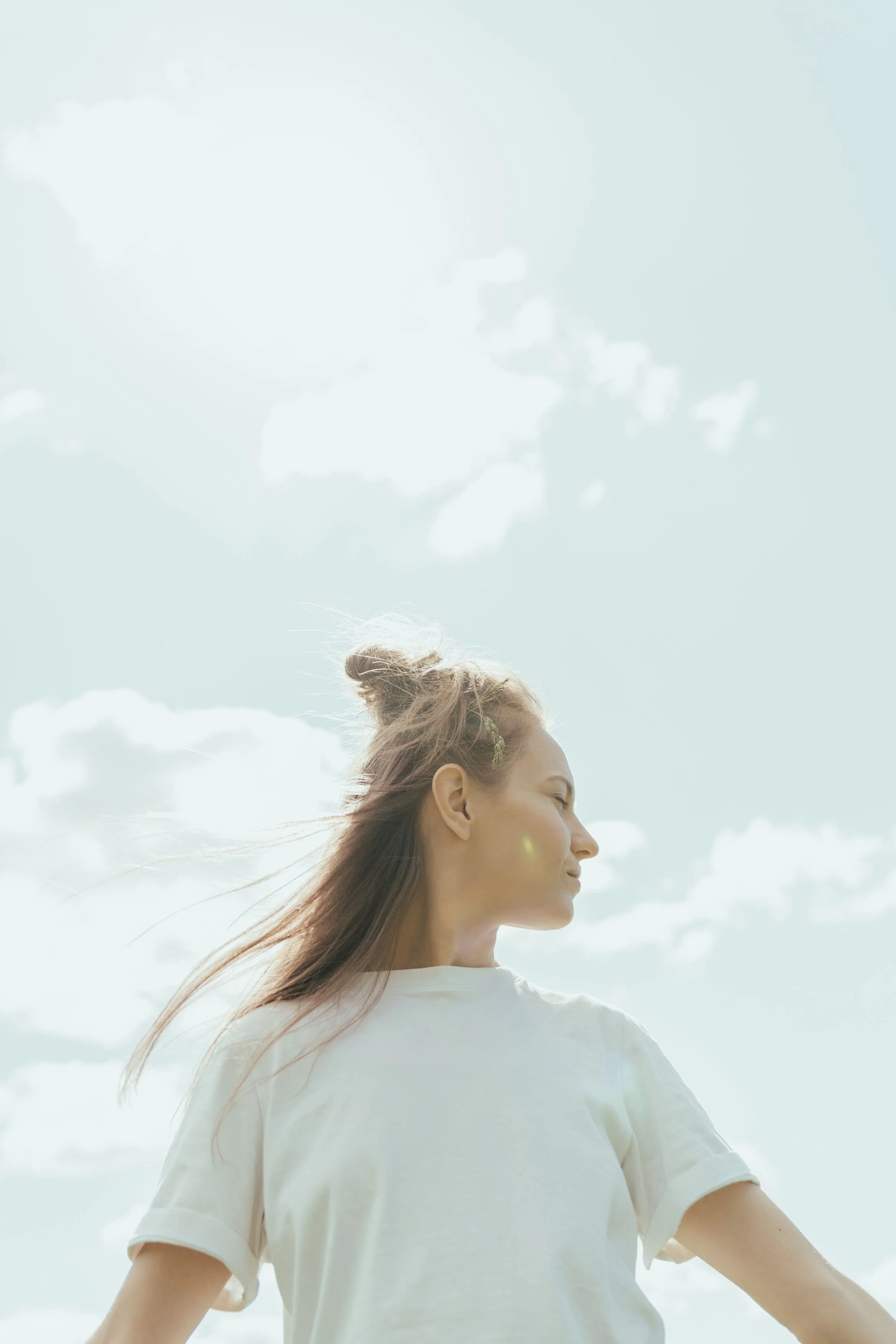 A woman with long hair in a bun and wearing a white T-shirt standing outdoors against a sky with scattered clouds.