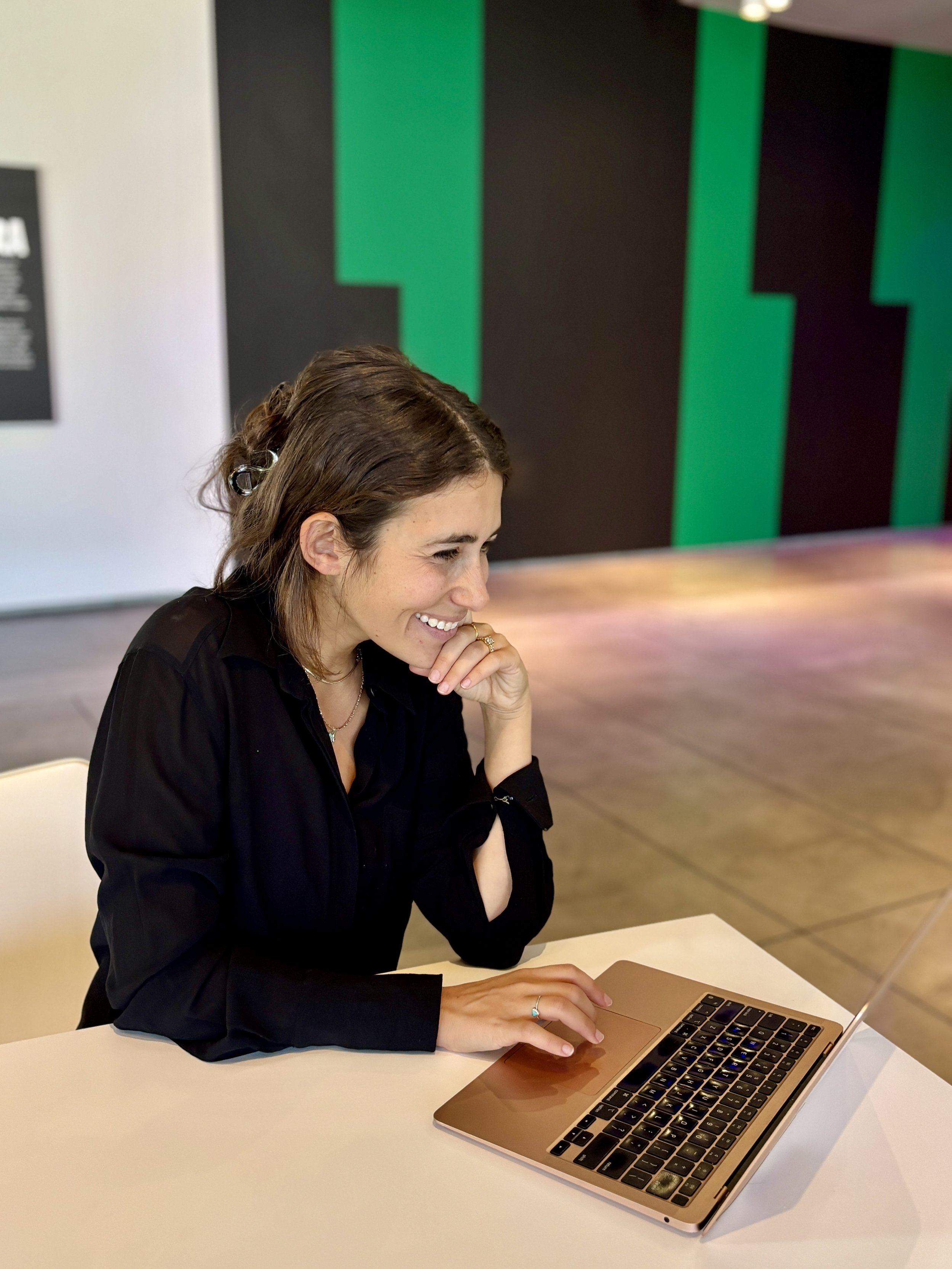 A woman with brown hair and a black shirt sitting at a white table, working on a gold-colored laptop, smiling and looking at the screen.