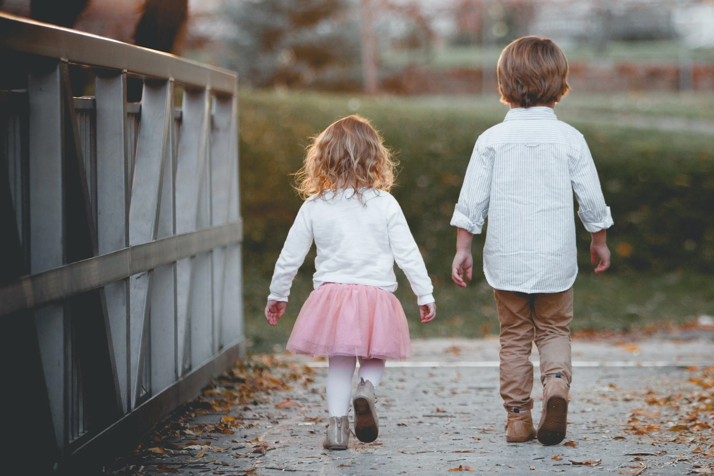 Two young children, a girl in a pink tutu, white tights, and beige boots, and a boy in tan pants and a striped shirt, walk hand in hand on a leaf-covered sidewalk in autumn.