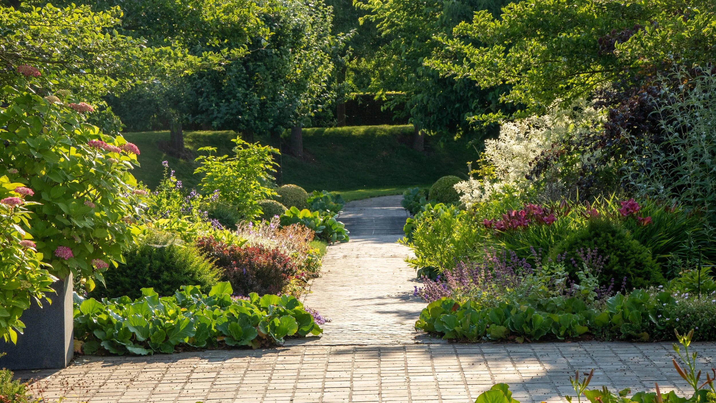 A garden pathway lined with colorful flowers and lush greenery, leading into a shaded area with trees.