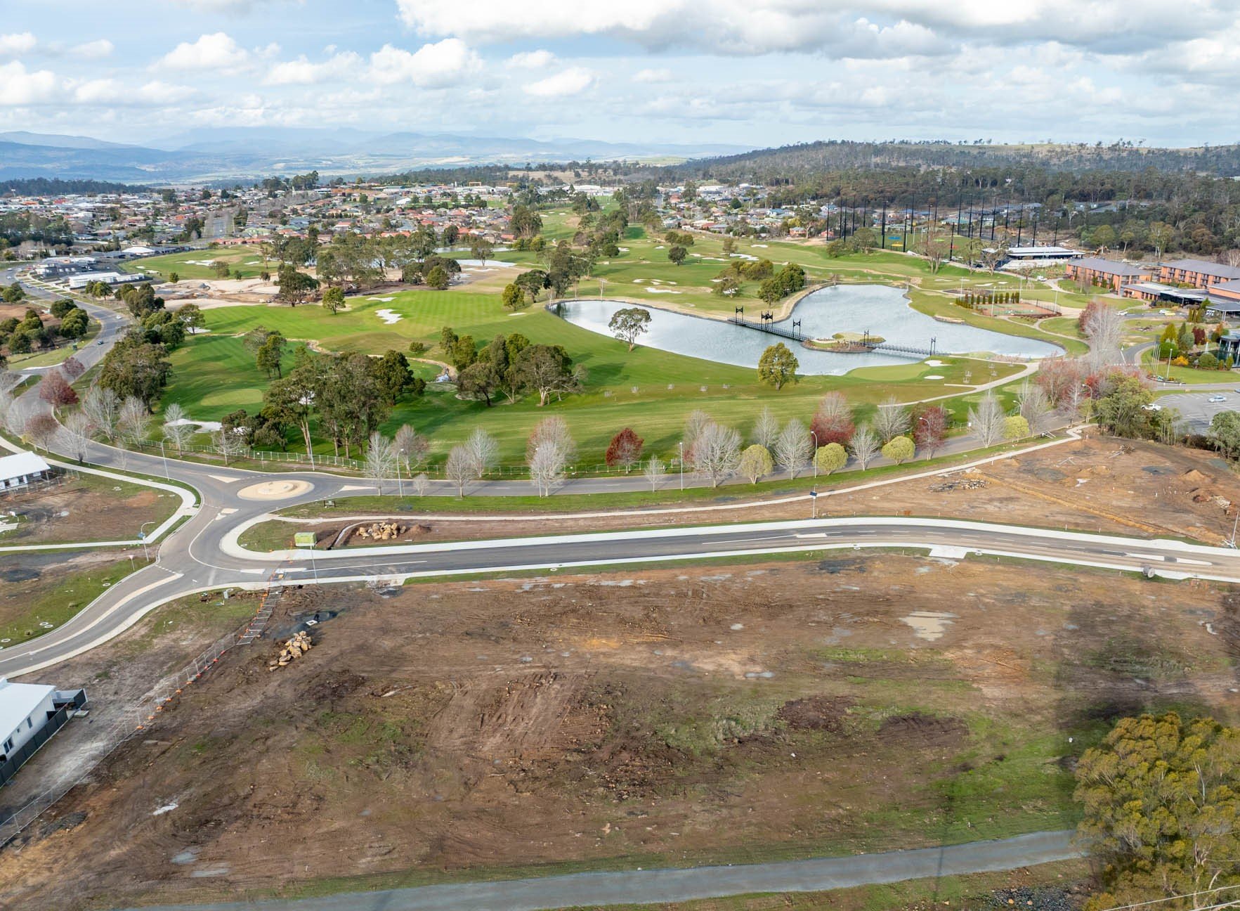 Aerial view of a golf course with ponds, trees, walking paths, surrounding buildings, and a nearby road under partly cloudy skies.