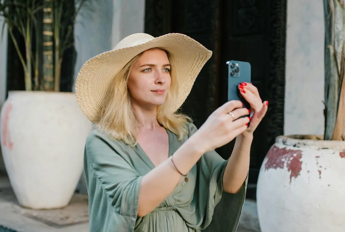 A woman wearing a wide-brimmed sunhat and a green dress taking a selfie with her smartphone outdoors.