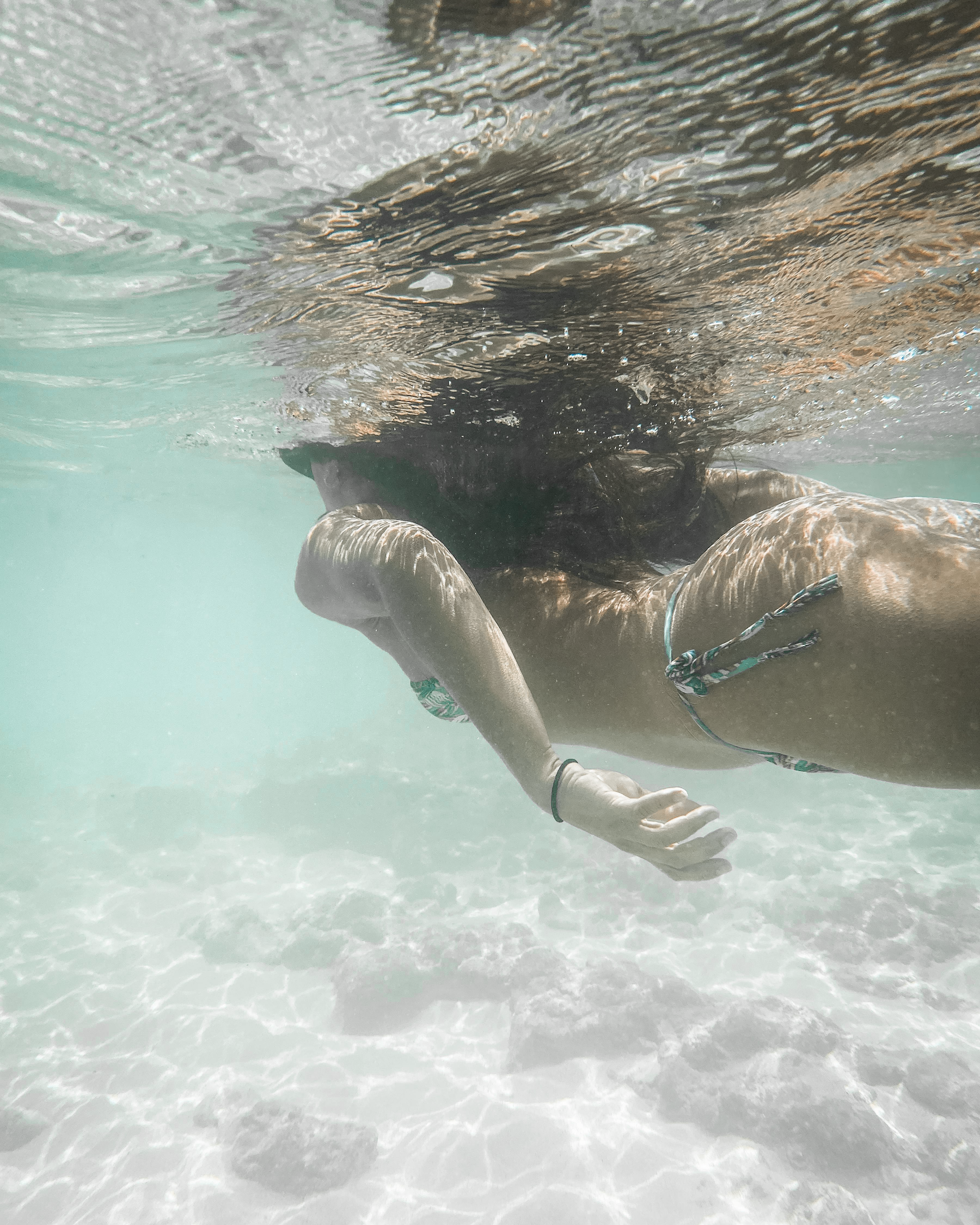 A woman swimming underwater in the ocean, wearing a bikini, with her arm bent and her hand near her face, and her hair floating freely.