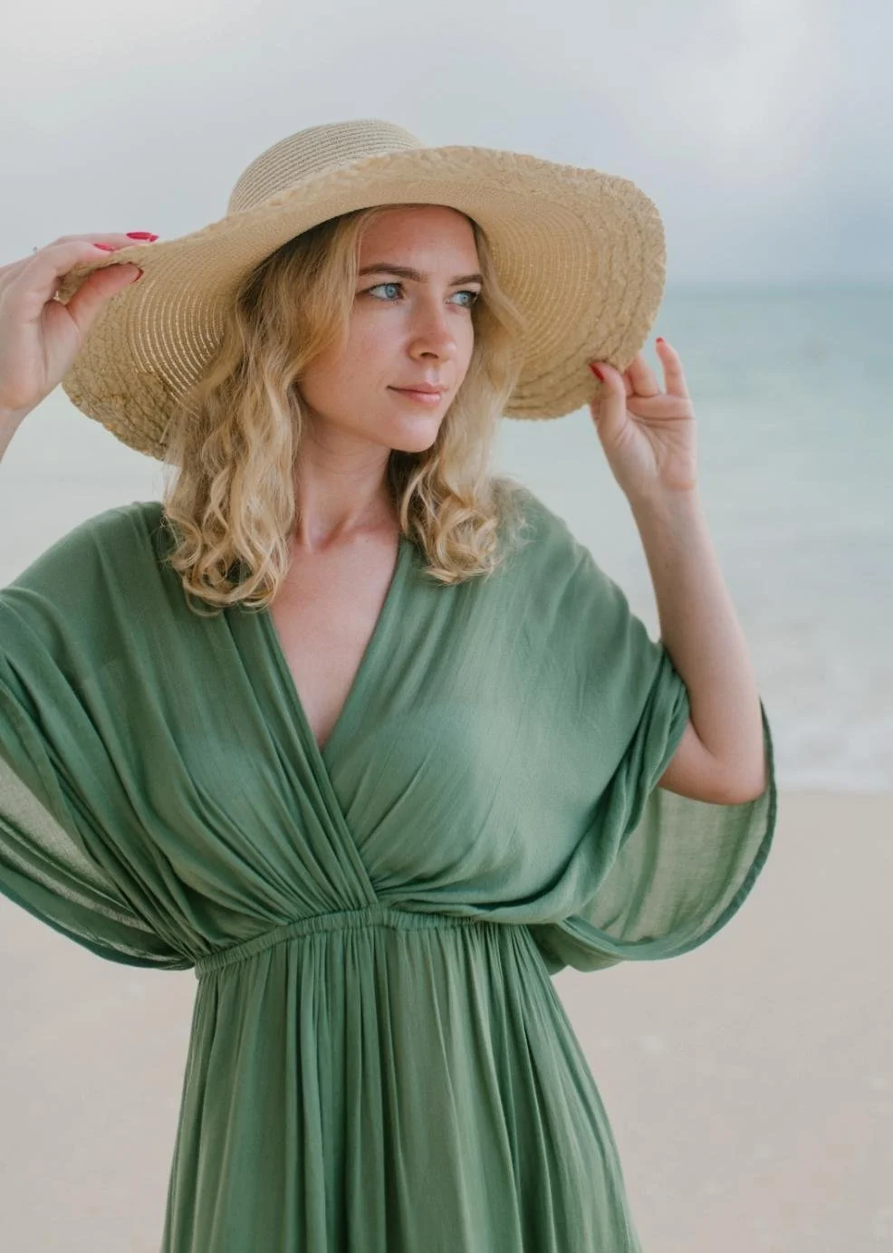 A woman in a green dress and wide-brimmed straw hat looks contemplatively at the beach, with the ocean and sky in the background.