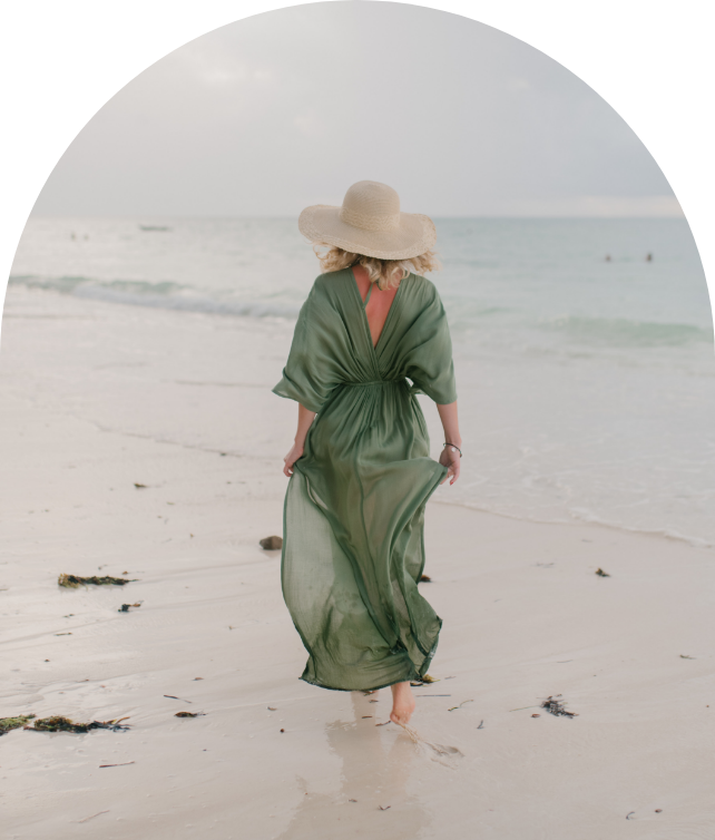 Woman in a green sheer dress and a wide-brimmed hat walking on a beach along the shoreline with ocean waves in the background.
