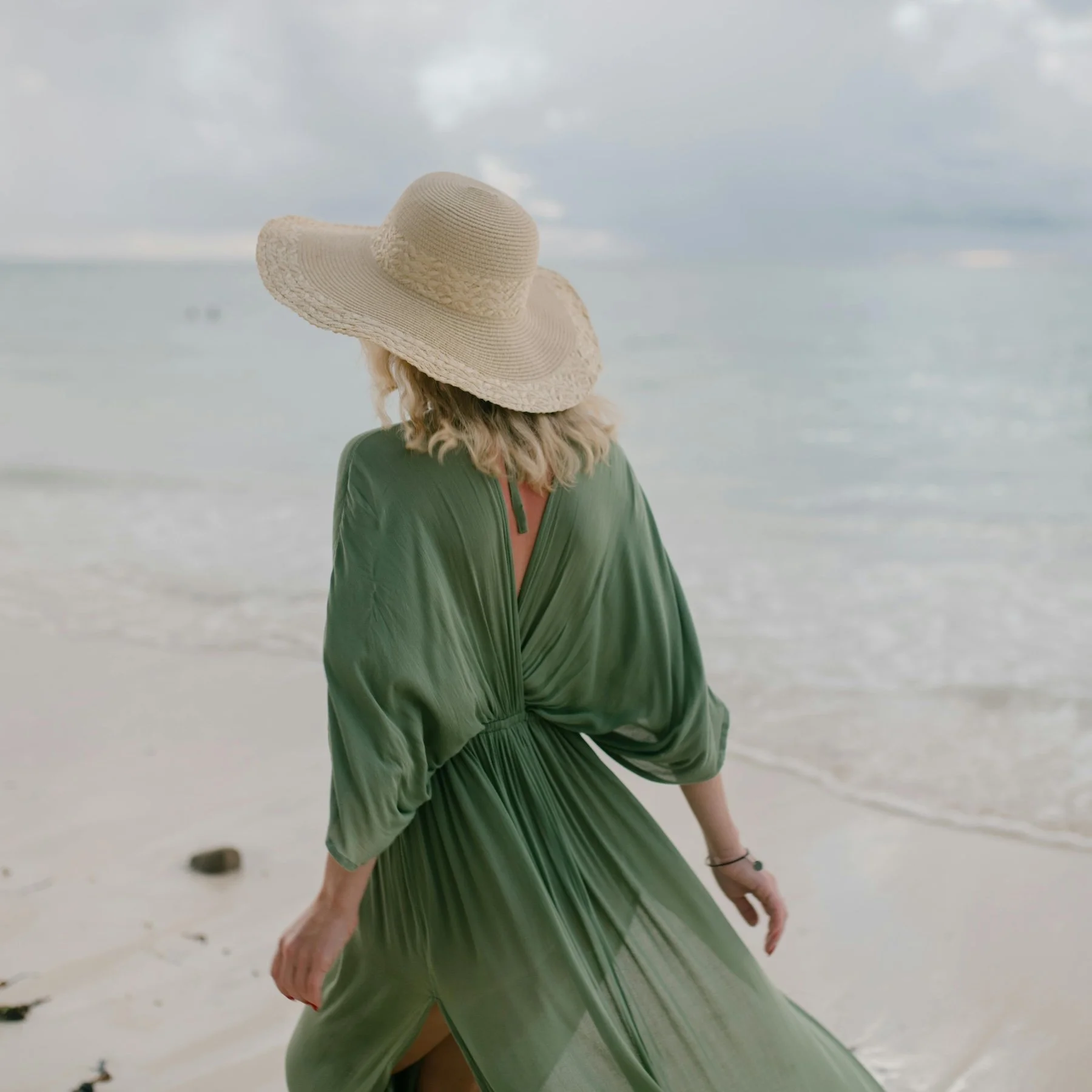 Woman wearing a large straw hat and a flowing green dress standing on a beach near the ocean.
