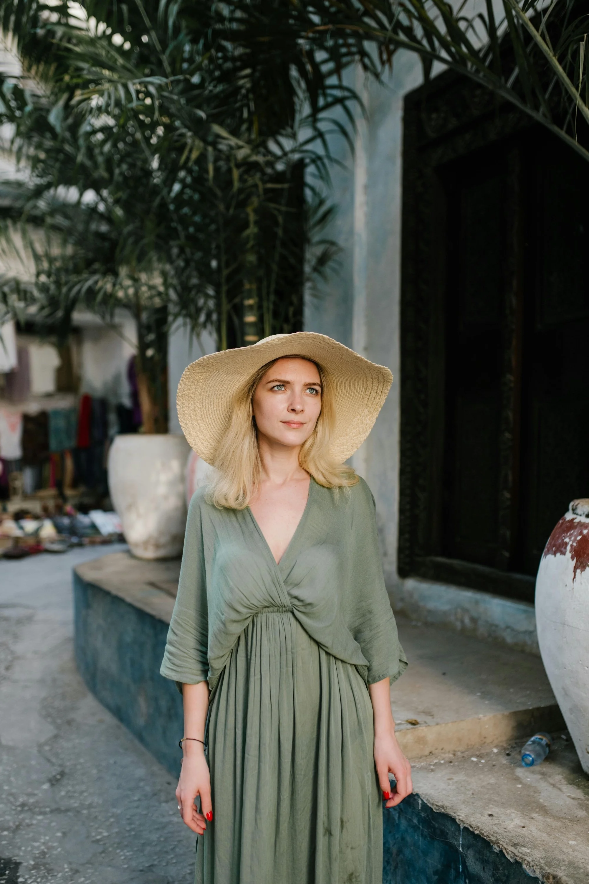 A woman in a green dress and wide-brimmed straw hat standing outdoors near large pots and lush greenery.