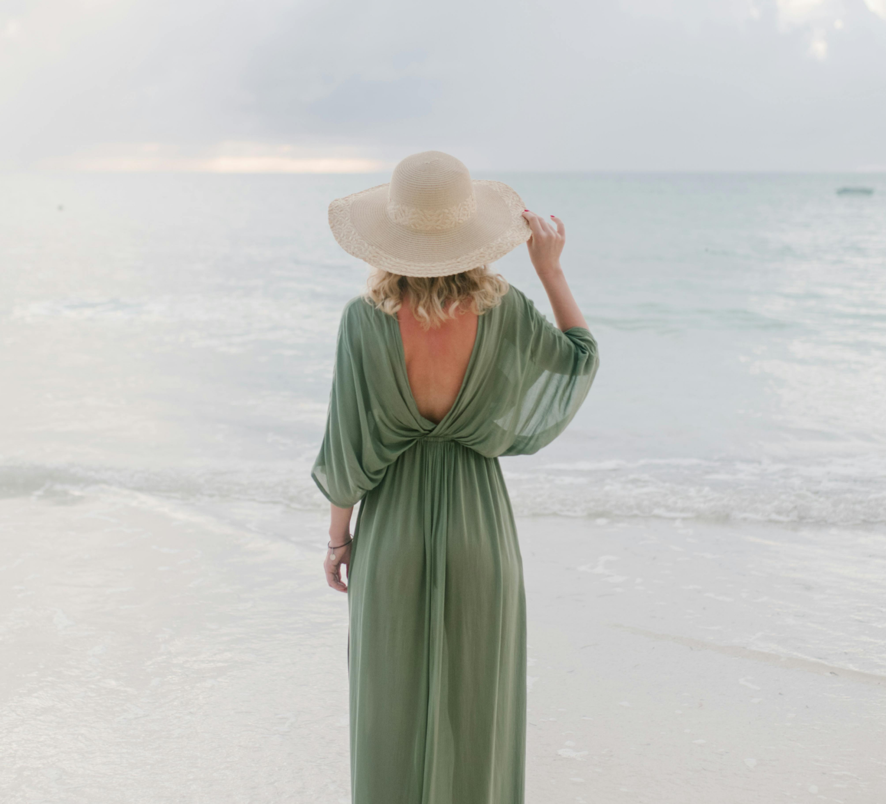Woman in a green dress and wide-brimmed hat standing on the beach, facing the ocean with her back.