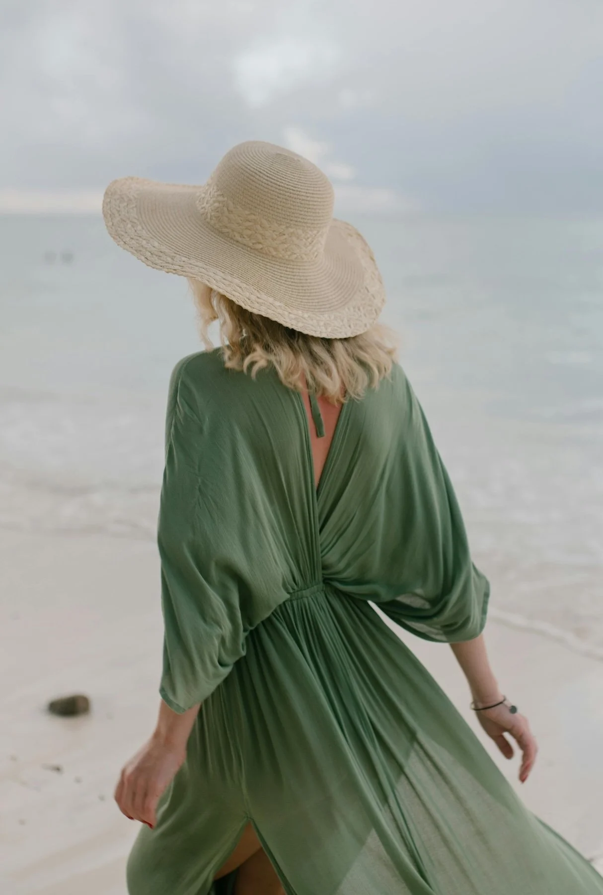 A woman wearing a large beige sunhat and a flowing green dress standing on a beach, facing the ocean.