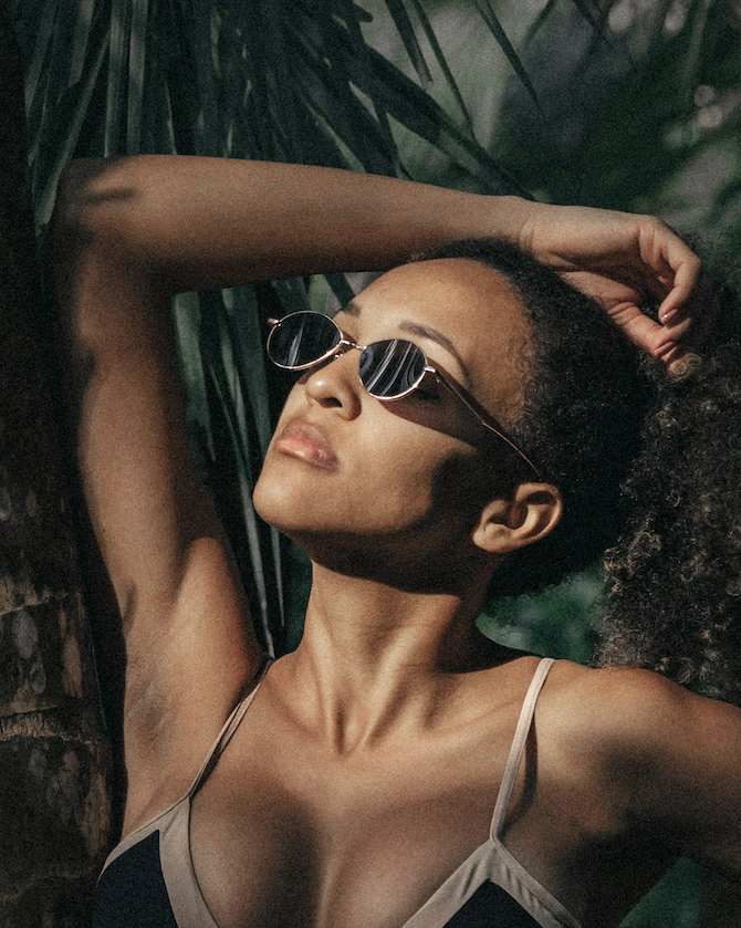 A woman with dark curly hair wearing sunglasses and a sleeveless top, leaning against a palm tree with her eyes closed in sunlight.