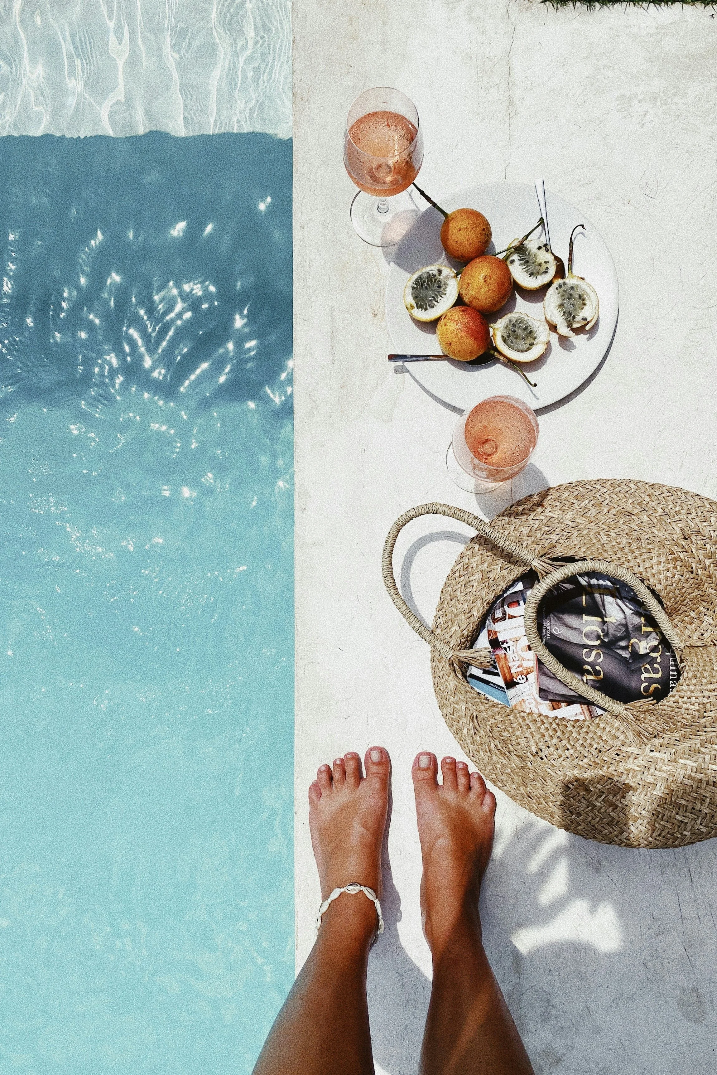 A view of a poolside scene with a person's feet in the foreground, a woven bag with magazines, a round white tray with three kiwifruits, blindfolded, and two glasses of pink wine on white surface next to a blue pool.