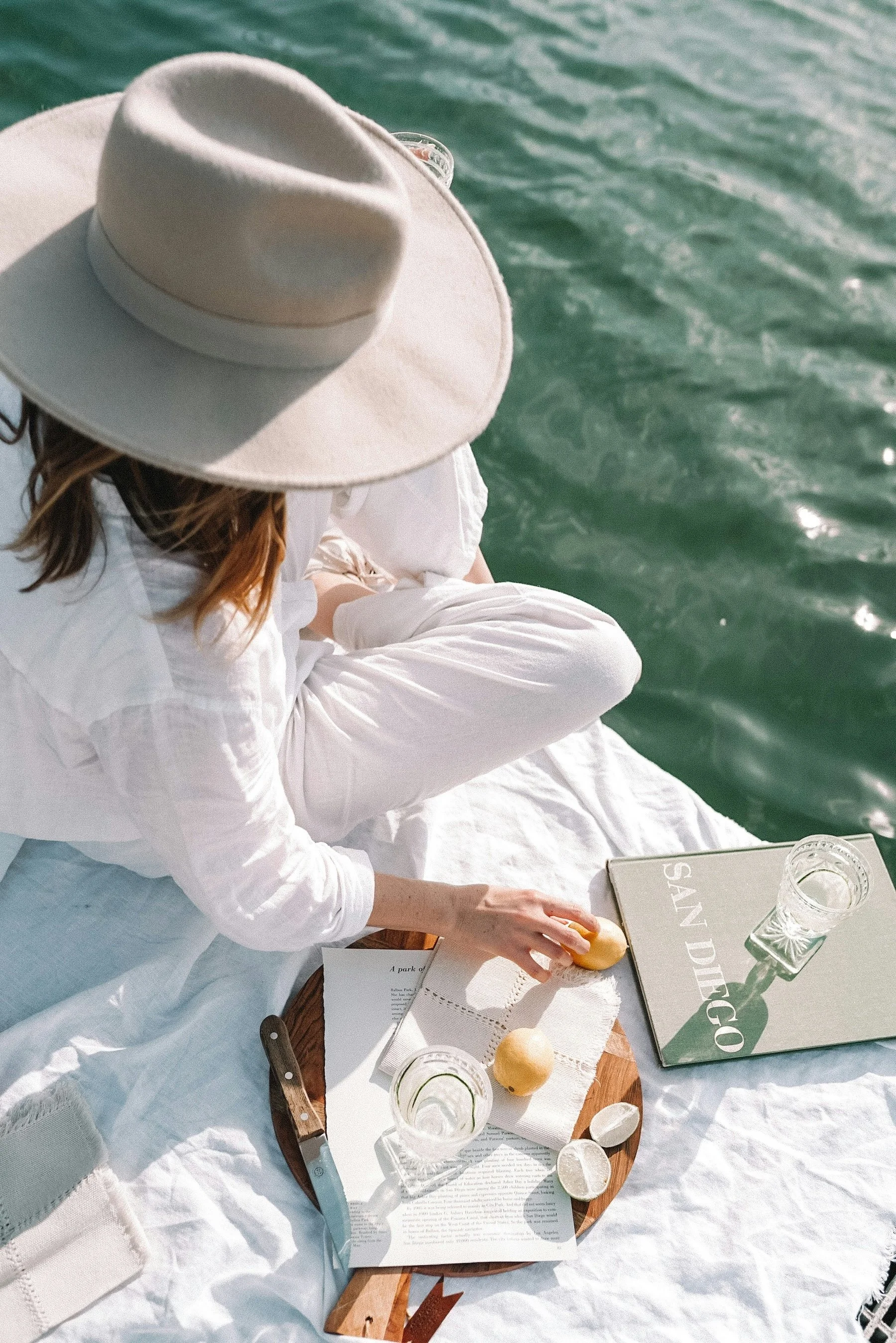 Woman in white sitting on a white blanket by the water, wearing a wide-brimmed hat, picking up a lemon from a wooden tray with other lemons, glasses of water, and a menu.