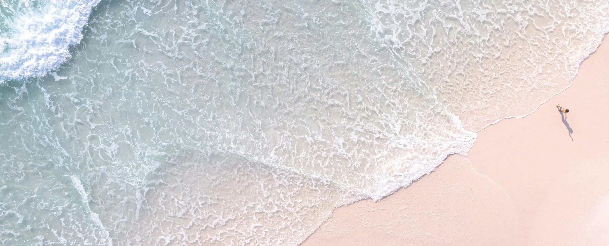 An aerial view of a person lying on the sandy beach near the shoreline with waves coming in.