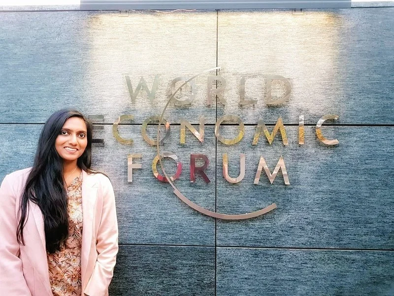 A woman with long black hair wearing a pink blazer and floral top, standing in front of a large metallic sign that reads 'World Economic Forum' on a dark blue tiled wall.