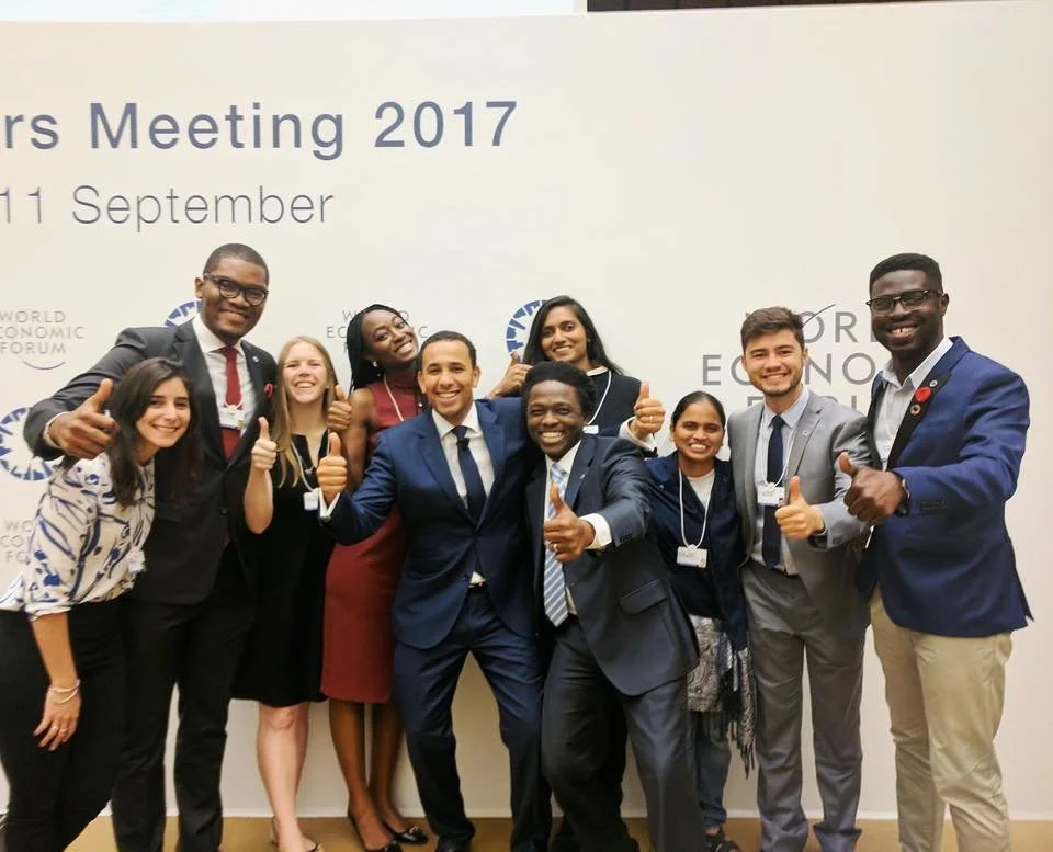 Group of nine diverse people in formal attire smiling and giving thumbs up at a conference, some with conference badges, in front of a backdrop that reads 'World Economic Forum'. Group includes men and women of different ethnicities.
