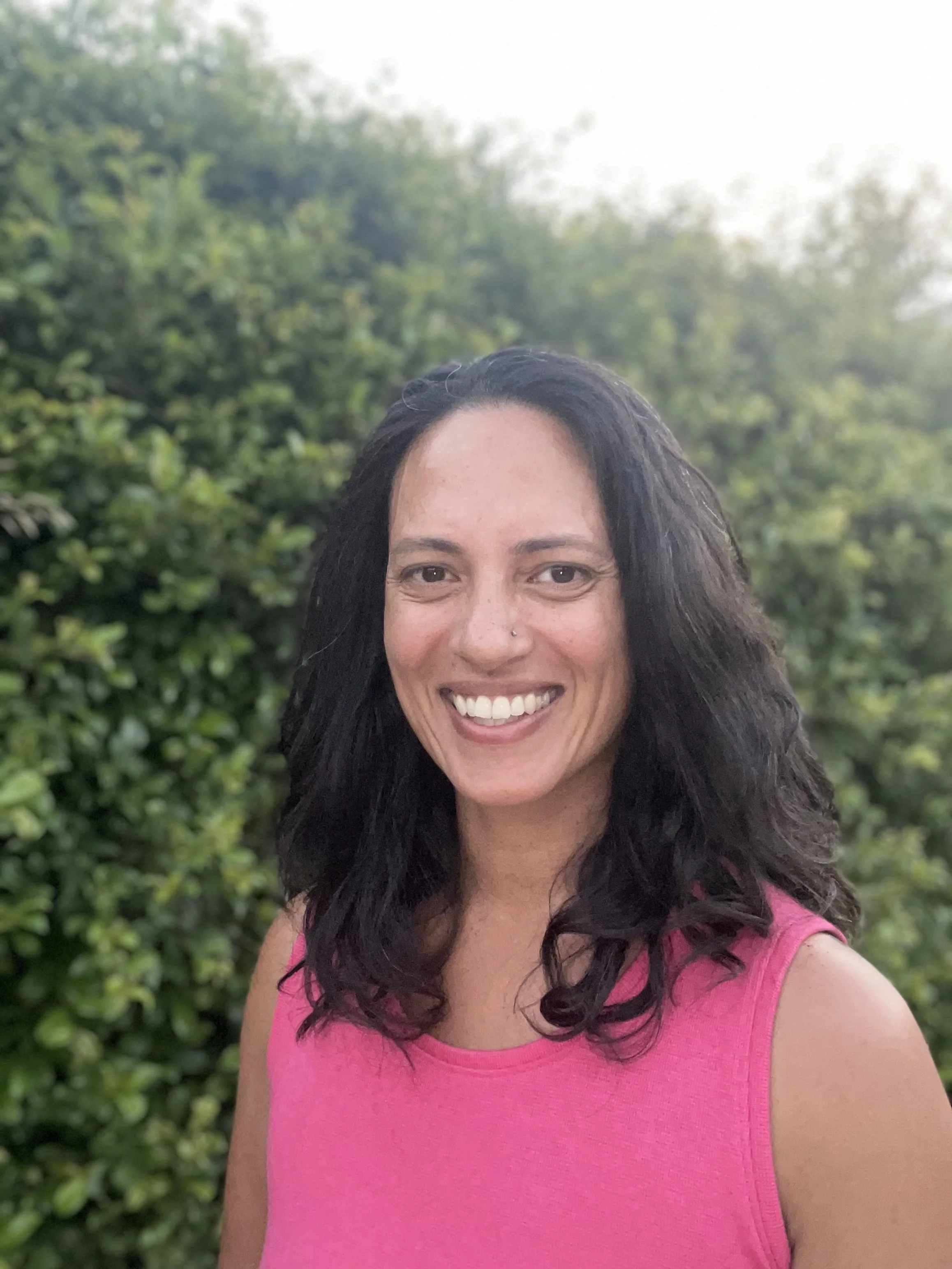 A smiling woman with black, shoulder-length, wavy hair, wearing a pink sleeveless top, standing outdoors with green bushes and trees in the background.