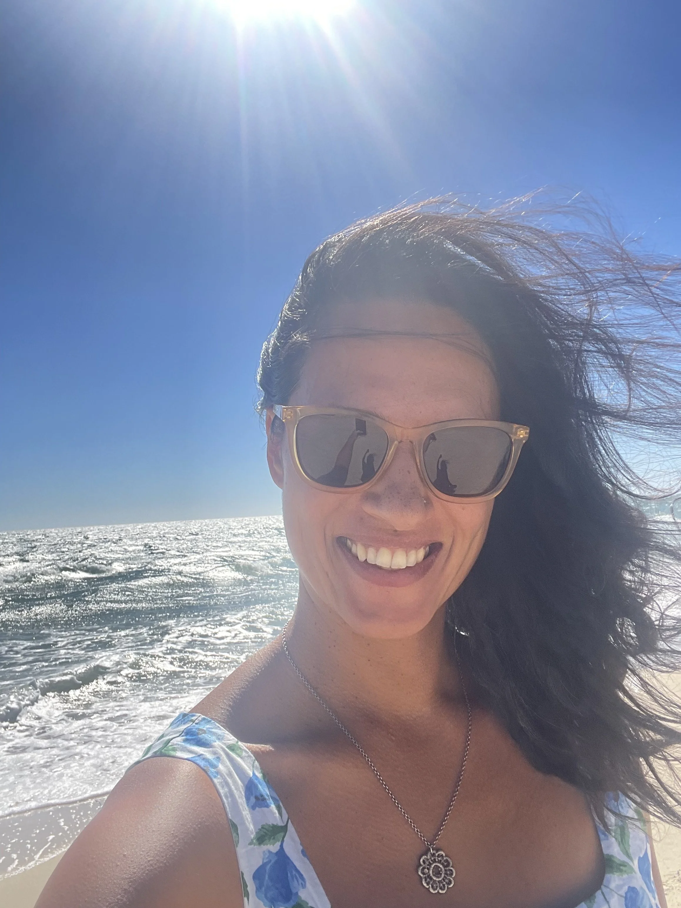 A woman smiling at the camera on a sunny beach, wearing sunglasses and a floral dress with ocean waves in the background.