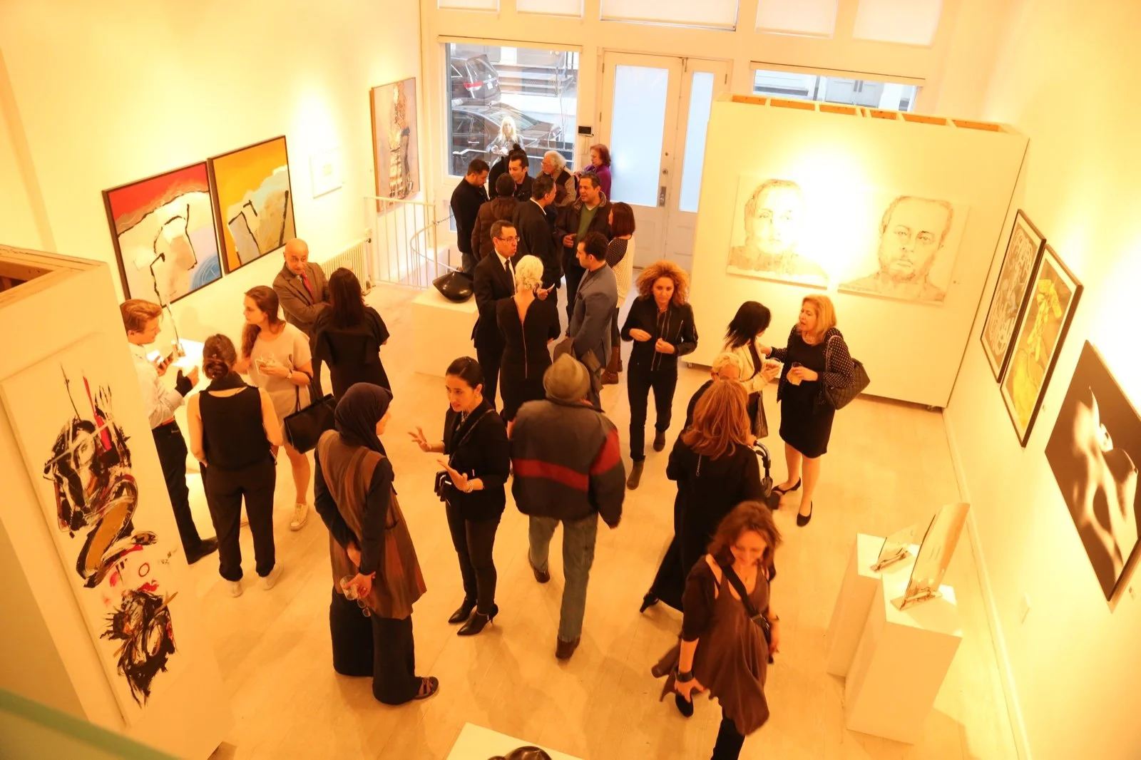 Two women talking in an art gallery, with colorful abstract paintings on the wall behind them. Other gallery visitors are visible in the background.