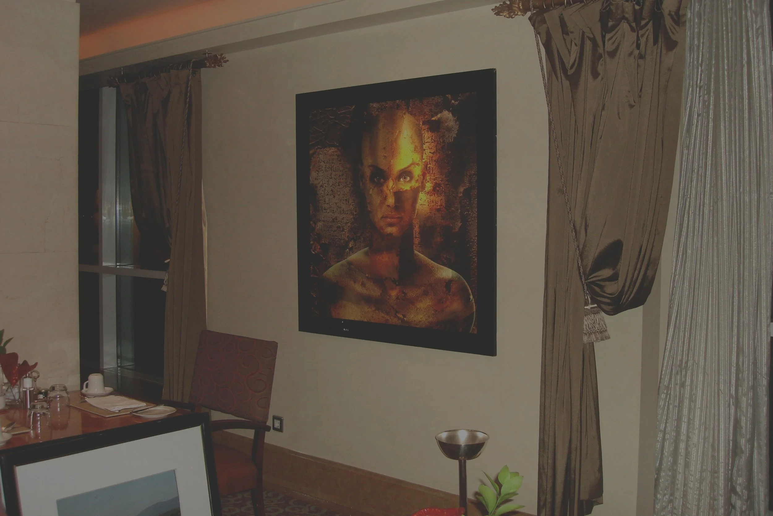A framed modern art portrait of a woman with an intense expression hanging on a beige wall in a restaurant or hotel room, with curtains, a table, and decor visible.