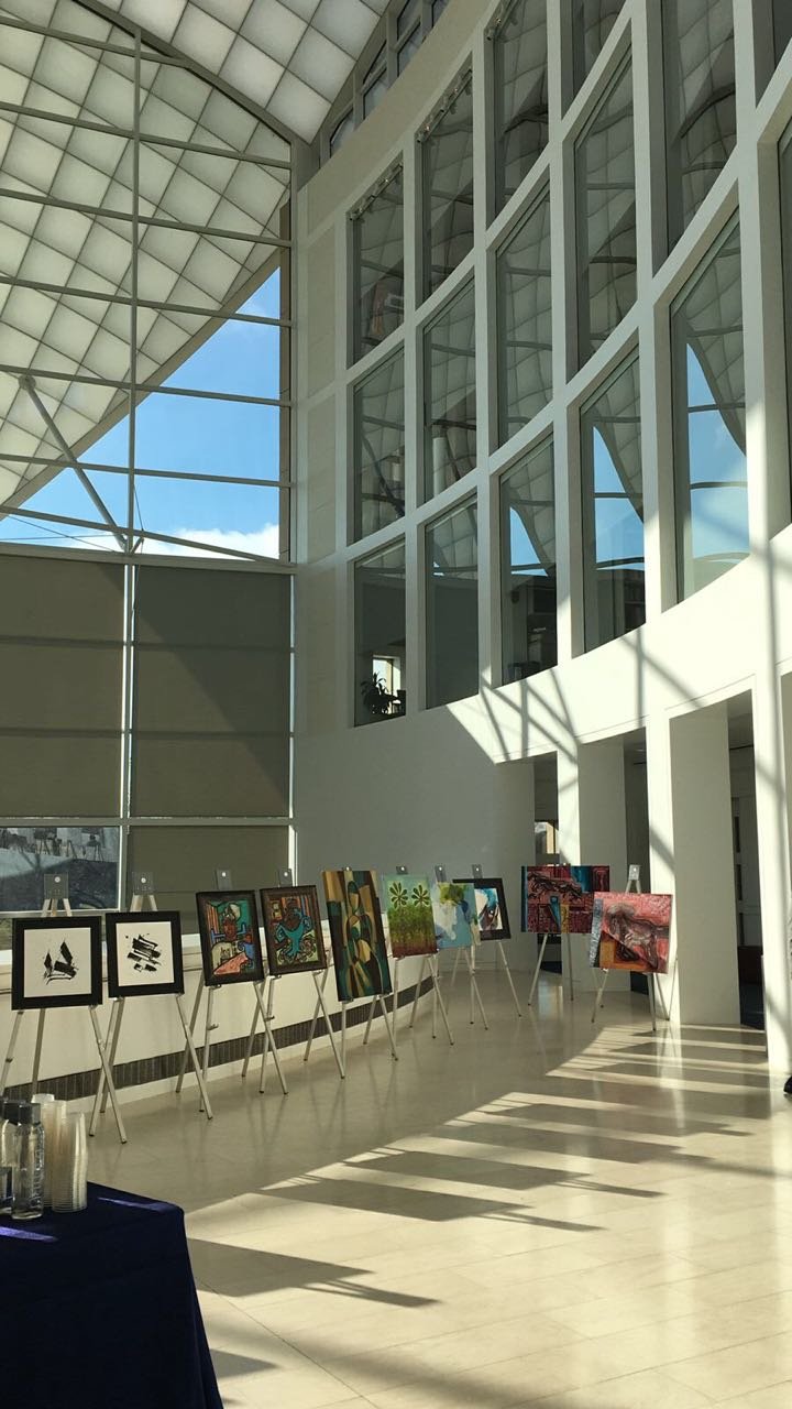 Art exhibit inside a modern building with large glass windows and shadow patterns on the floor, displaying colorful artwork on easels.