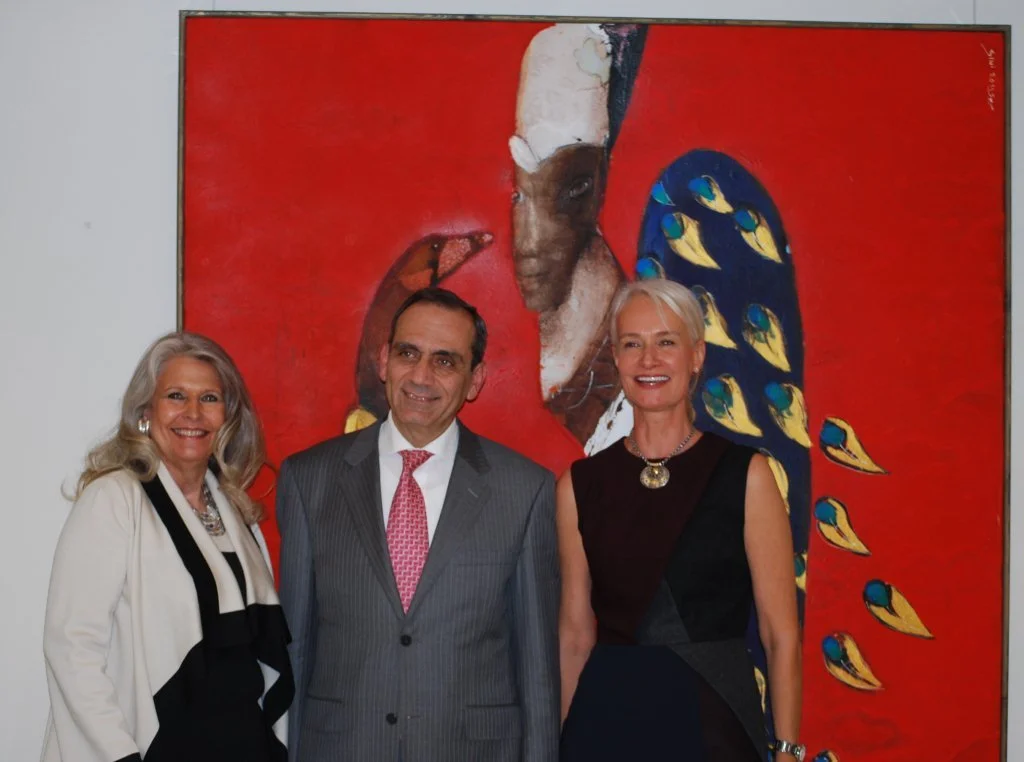Three people standing in front of a colorful abstract painting with a red background, a face, and peacock feather motifs.