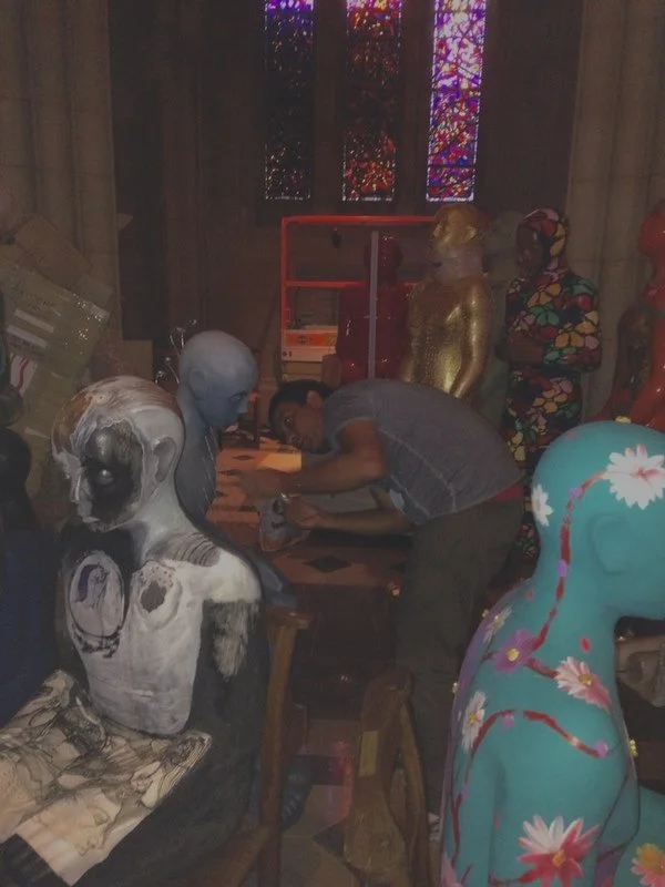 A man working on a sculpture in an art studio filled with colorful and metallic sculptures, including a dog figure with blue skin and a dog sculpture painted with flowers.