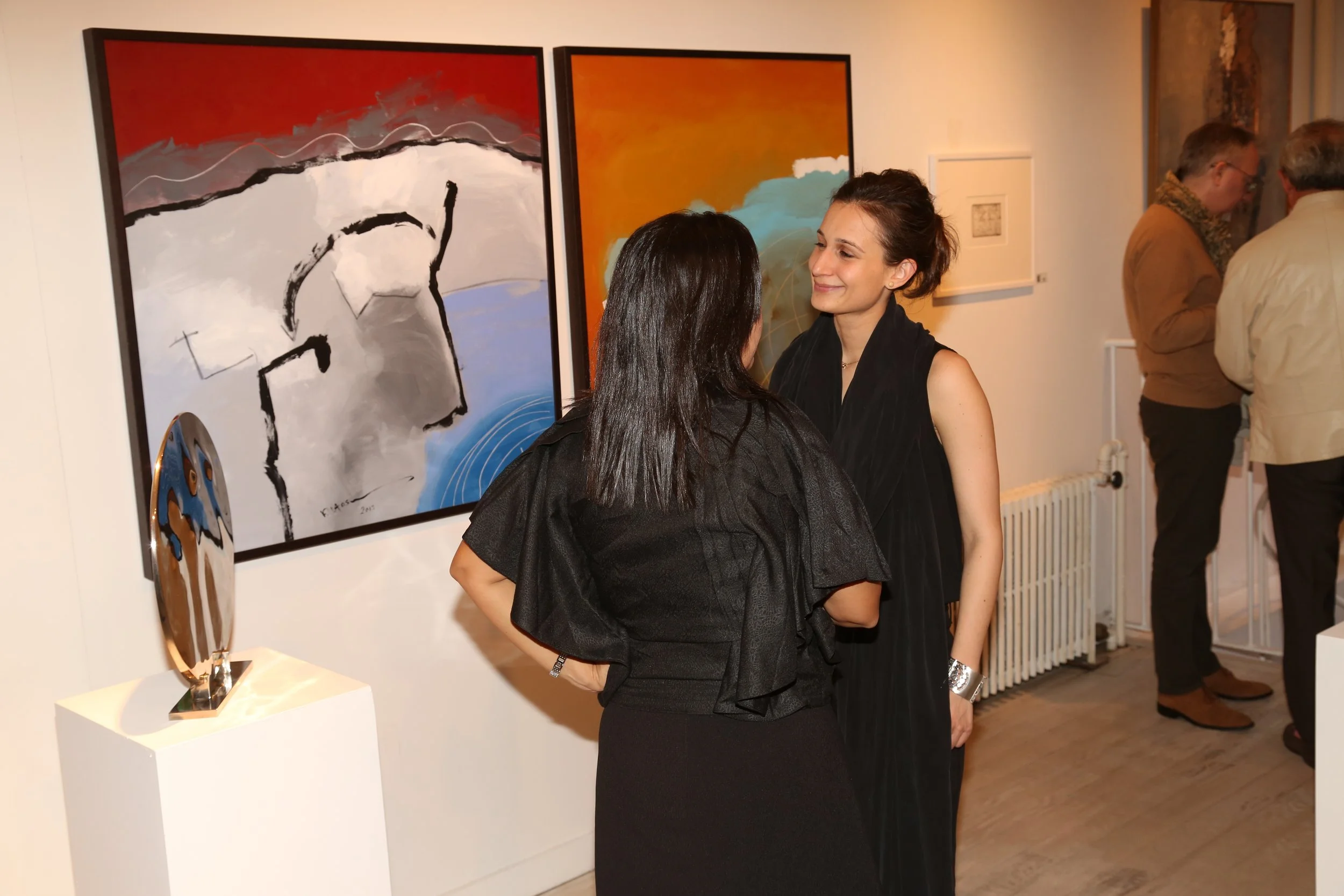 Two women talking in an art gallery, with colorful abstract paintings on the wall behind them. Other gallery visitors are visible in the background.