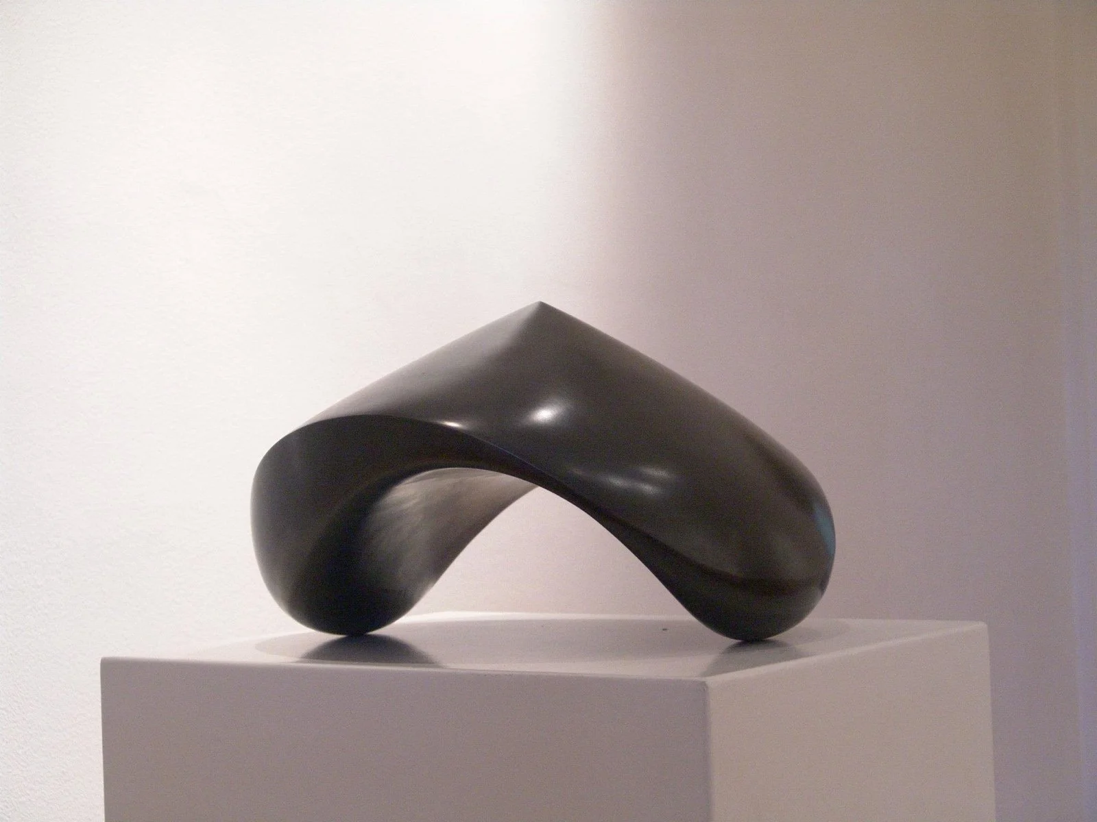 Abstract black sculpture with curved, flowing form on a white pedestal in a gallery setting.
