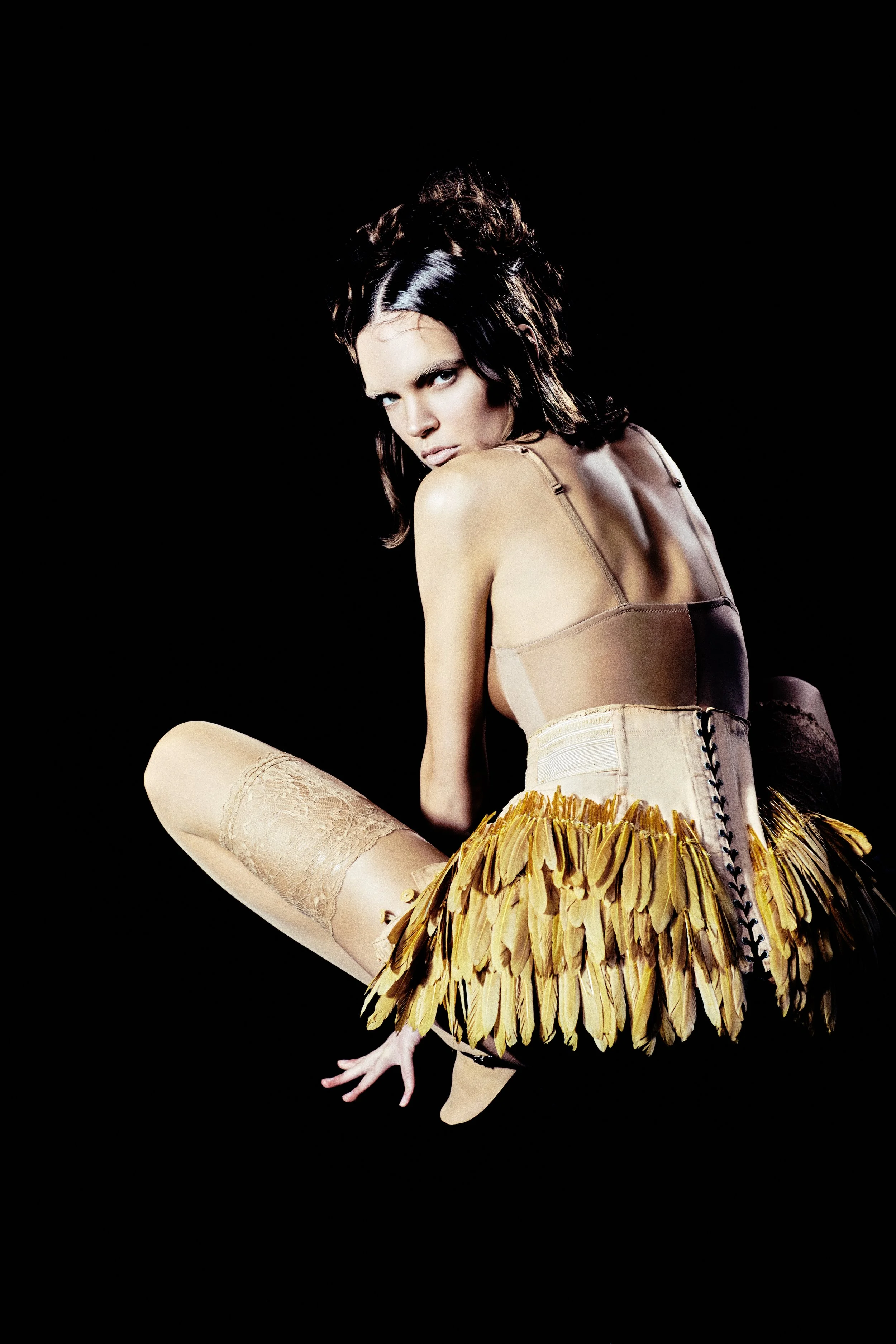 A woman with dark hair in a dramatic pose wearing a beige corset, beige stockings, and a yellow feathered skirt against a black background.