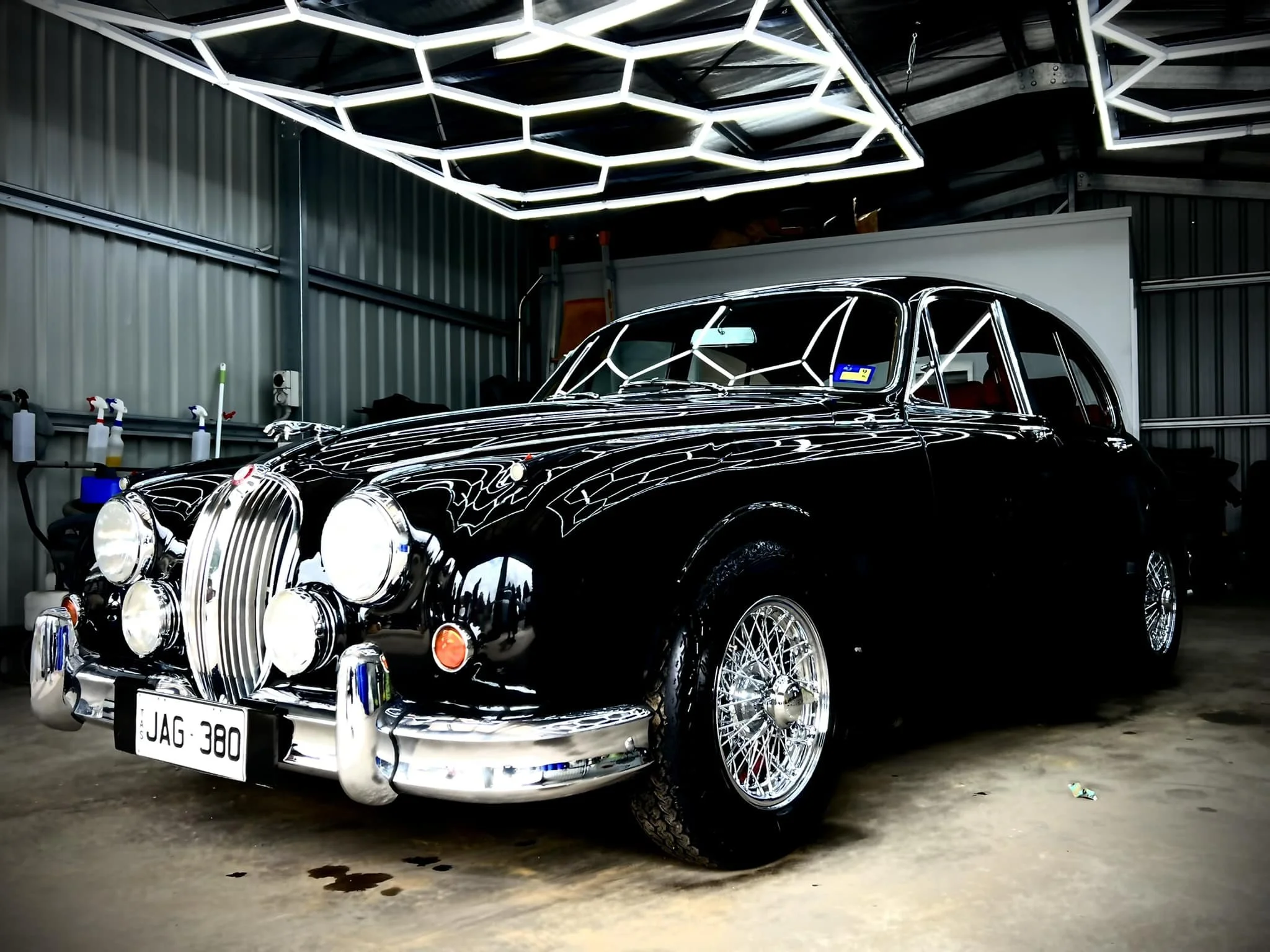 A classic black Jaguar vintage car in a garage with illuminated hexagon ceiling lights and a metal wall background.