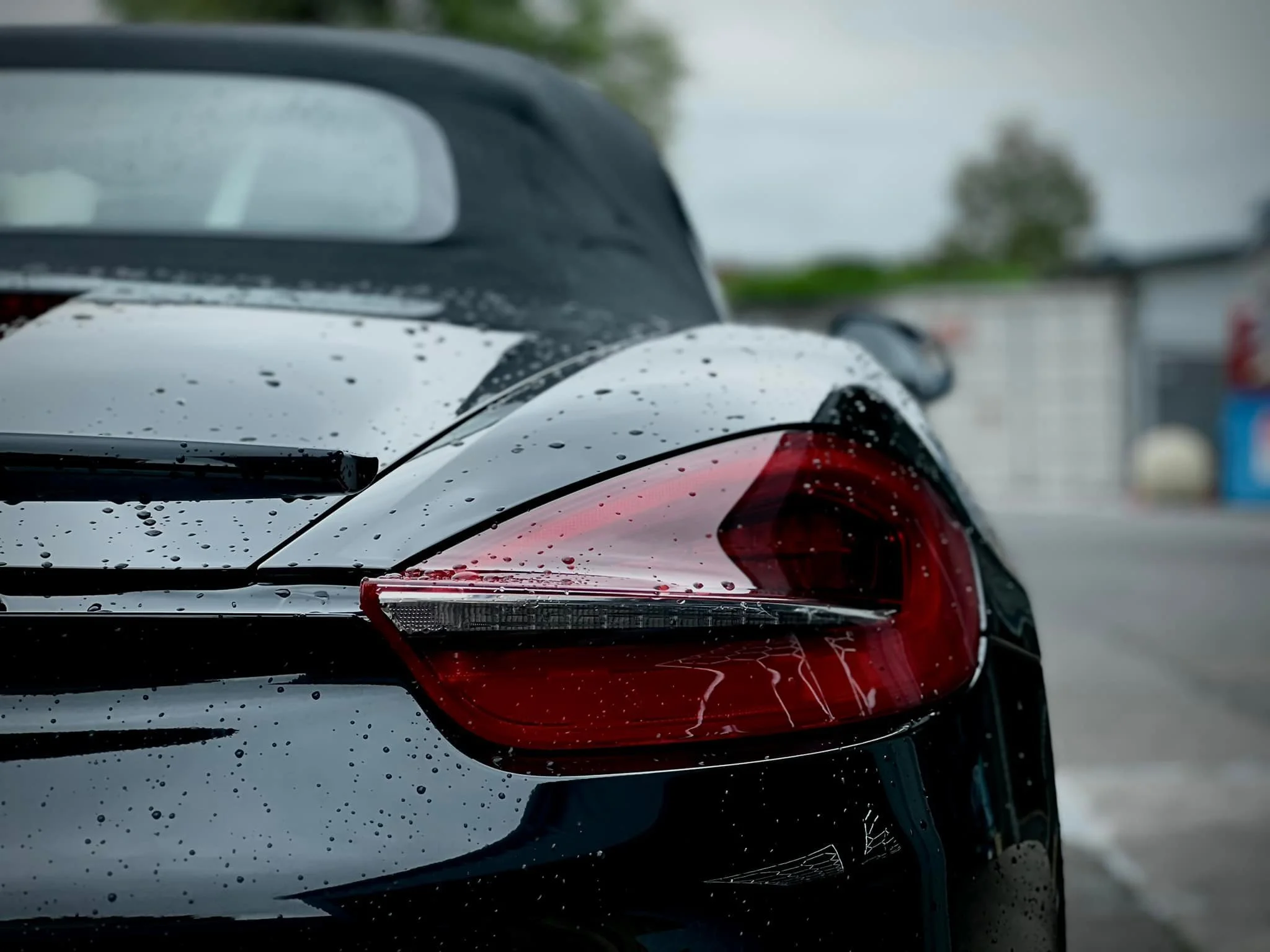 Close-up of the rear of a black sports car with water droplets and red taillights, on a rainy day.