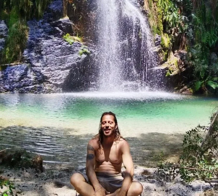 A man with long hair and a beard sitting on the sand in front of a waterfall and a clear pool of water surrounded by lush greenery.