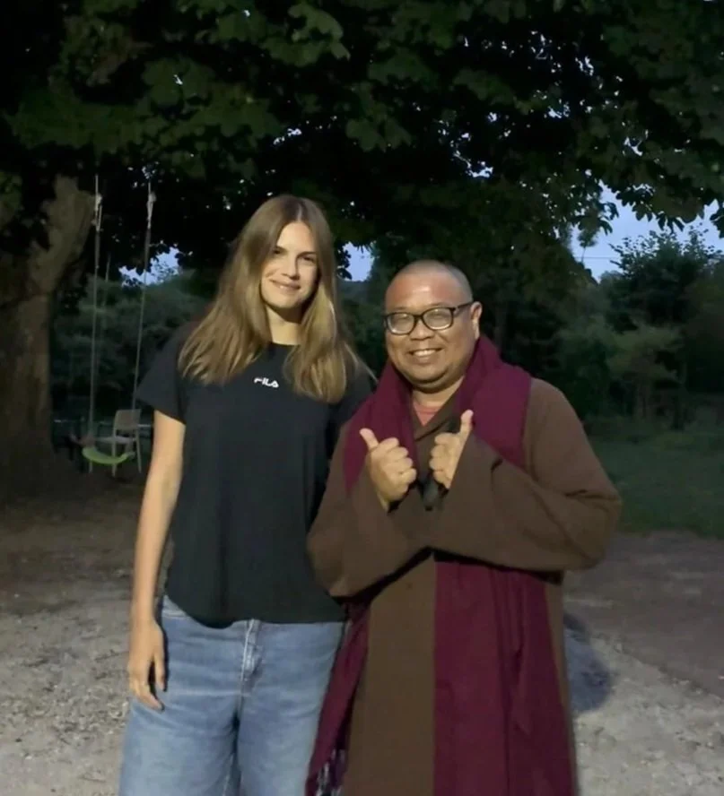 Two people smiling outdoors near trees, one young woman with long hair in a black T-shirt and jeans, and one man in glasses, wearing a brown coat and a maroon scarf, making a thumbs-up gesture.