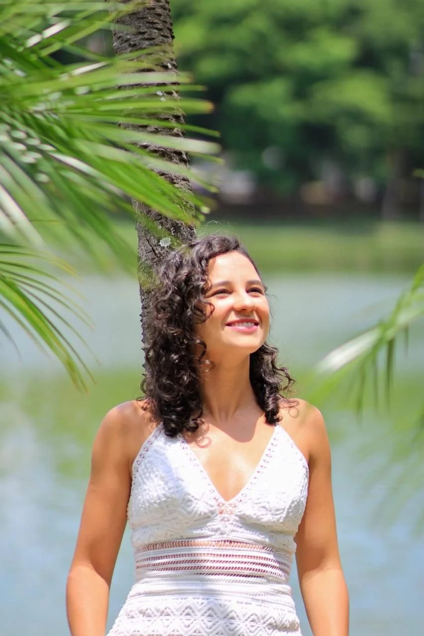 A woman with curly dark hair smiling outdoors near a body of water, surrounded by green trees and palm leaves.