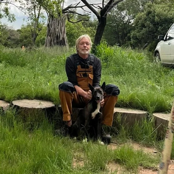 A man sits on a cut tree stump outdoors with a black dog, in a grassy area with trees and a white vehicle in the background.