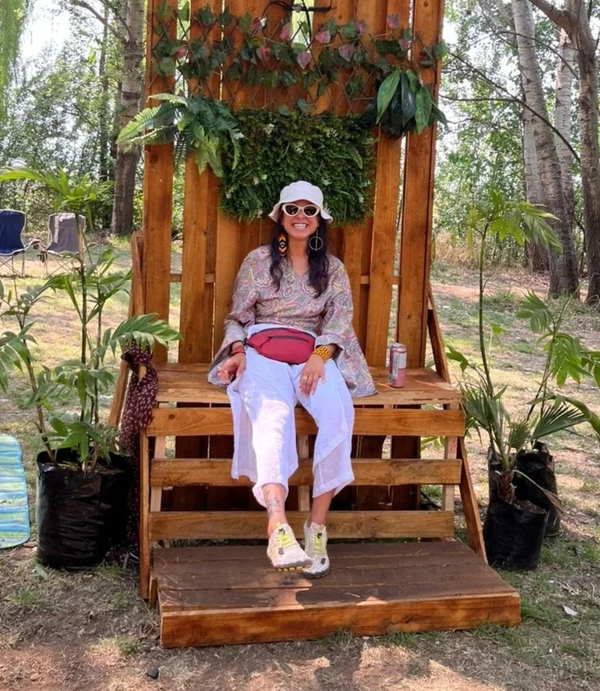 A woman sitting on a wooden throne-like chair outdoors, surrounded by plants and trees. She is smiling, wearing a white hat, sunglasses, a patterned long-sleeve top, white pants, and sneakers. There is a can on the right side of the chair and leafy decorations on the backrest.