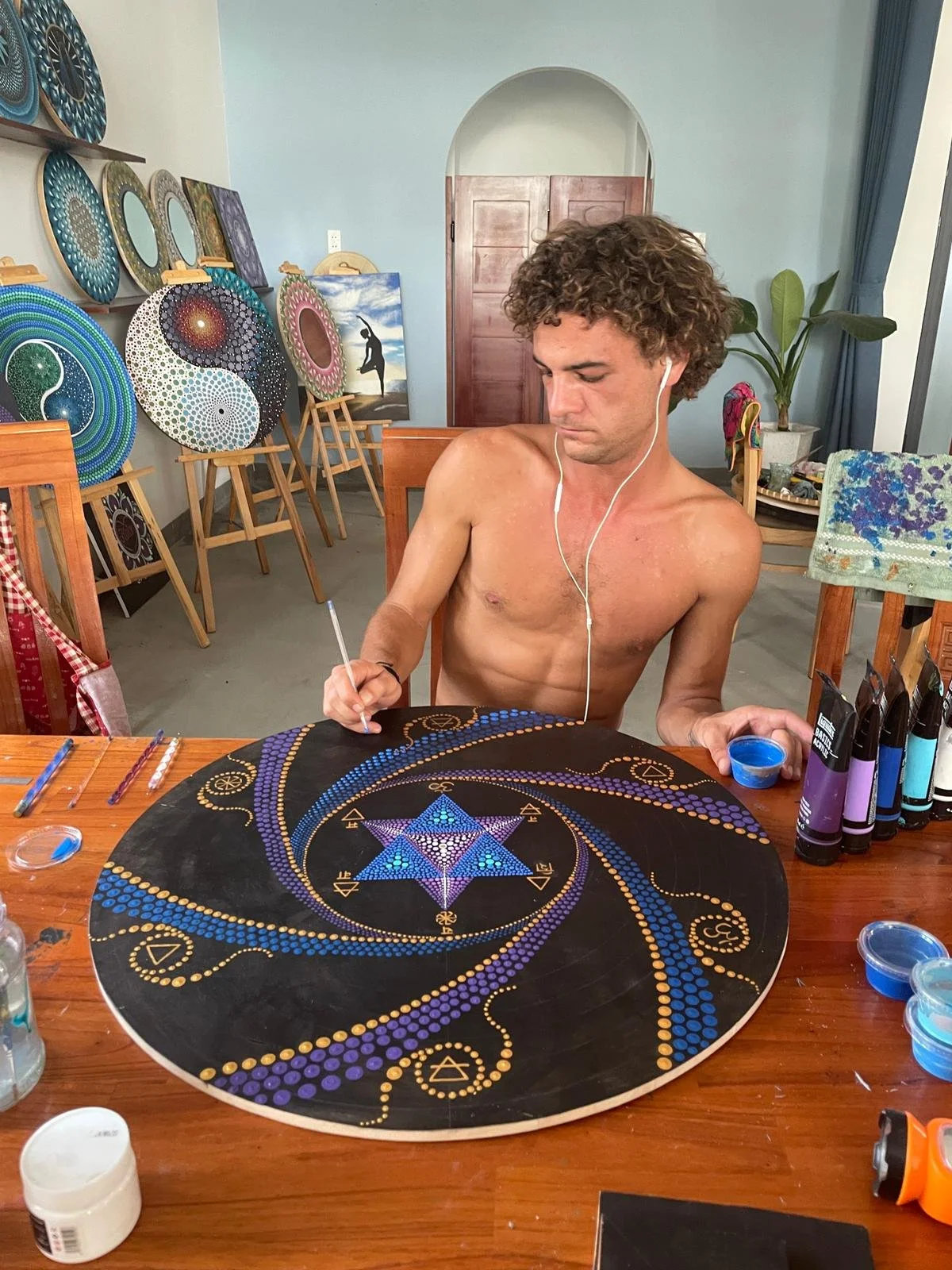 Julien Van Niekerk - A shirtless man with curly hair is creating a dot painting on a round black canvas, surrounded by paint containers and art supplies, with multiple colorful dot artworks displayed in the background.