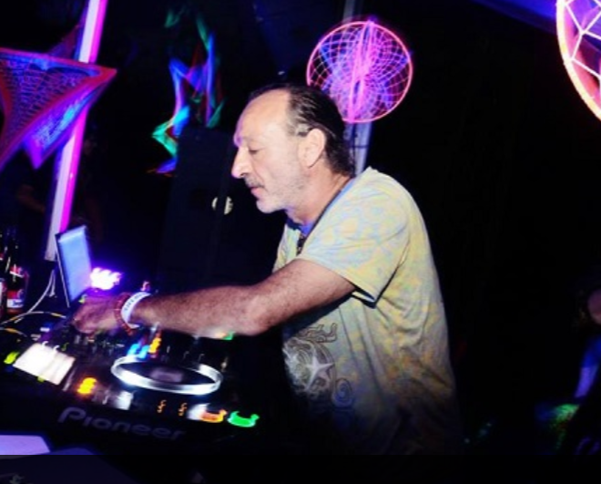 A DJ with short gray hair and a mustache, wearing a light-colored shirt, operating a Pioneer DJ controller in a club with neon lights and geometric designs in the background.