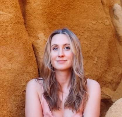 A woman with long blonde hair and a pink top, standing in front of a rocky, desert-like background.