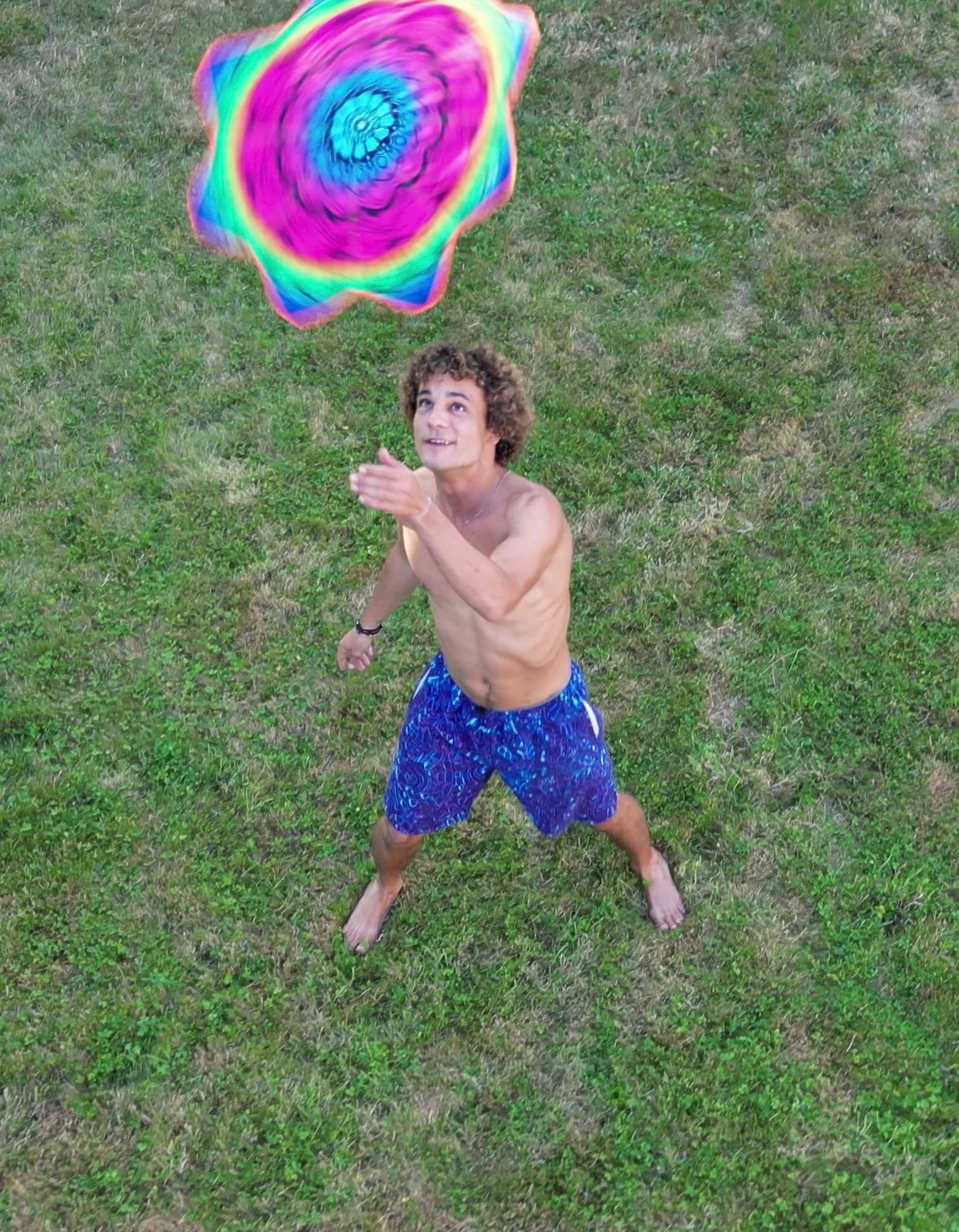Julien Van Niekerk - A shirtless young man with curly hair in blue shorts tossing a colorful, swirling, rainbow-patterned frisbee on a grassy lawn.