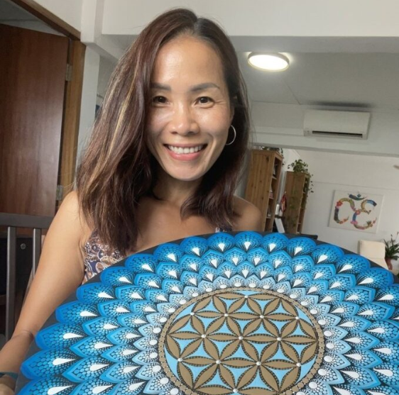 Smiling woman with shoulder-length brown hair holding a decorative blue and gold circular art piece in a home interior.