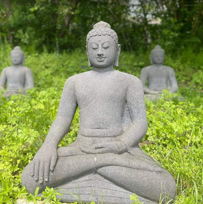 Three stone Buddha statues sitting in a green outdoor setting with lush foliage.