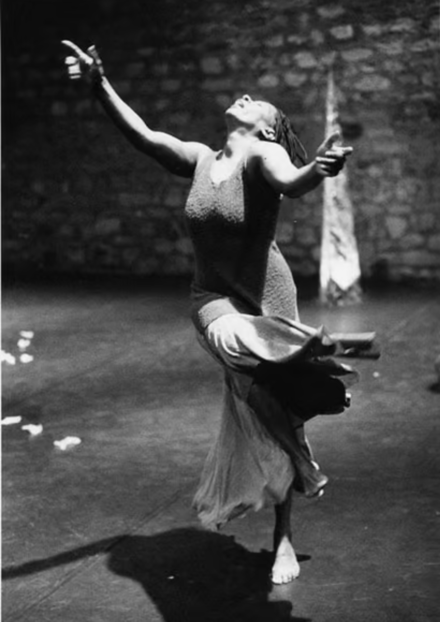 Une femme danse sur scène, vêtue d'une robe longue, avec un mur de briques en arrière-plan. Elle a les bras levés et le visage tourné vers le haut.