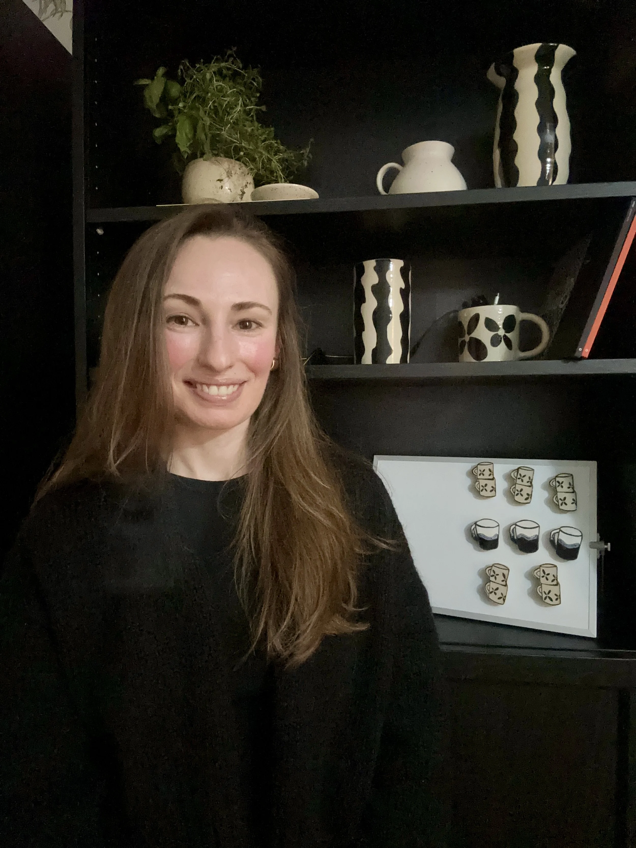 Portrait of Sarah, the ceramic artist behind La Céramique by Sarah, smiling in front of a black shelf filled with her handmade pottery.